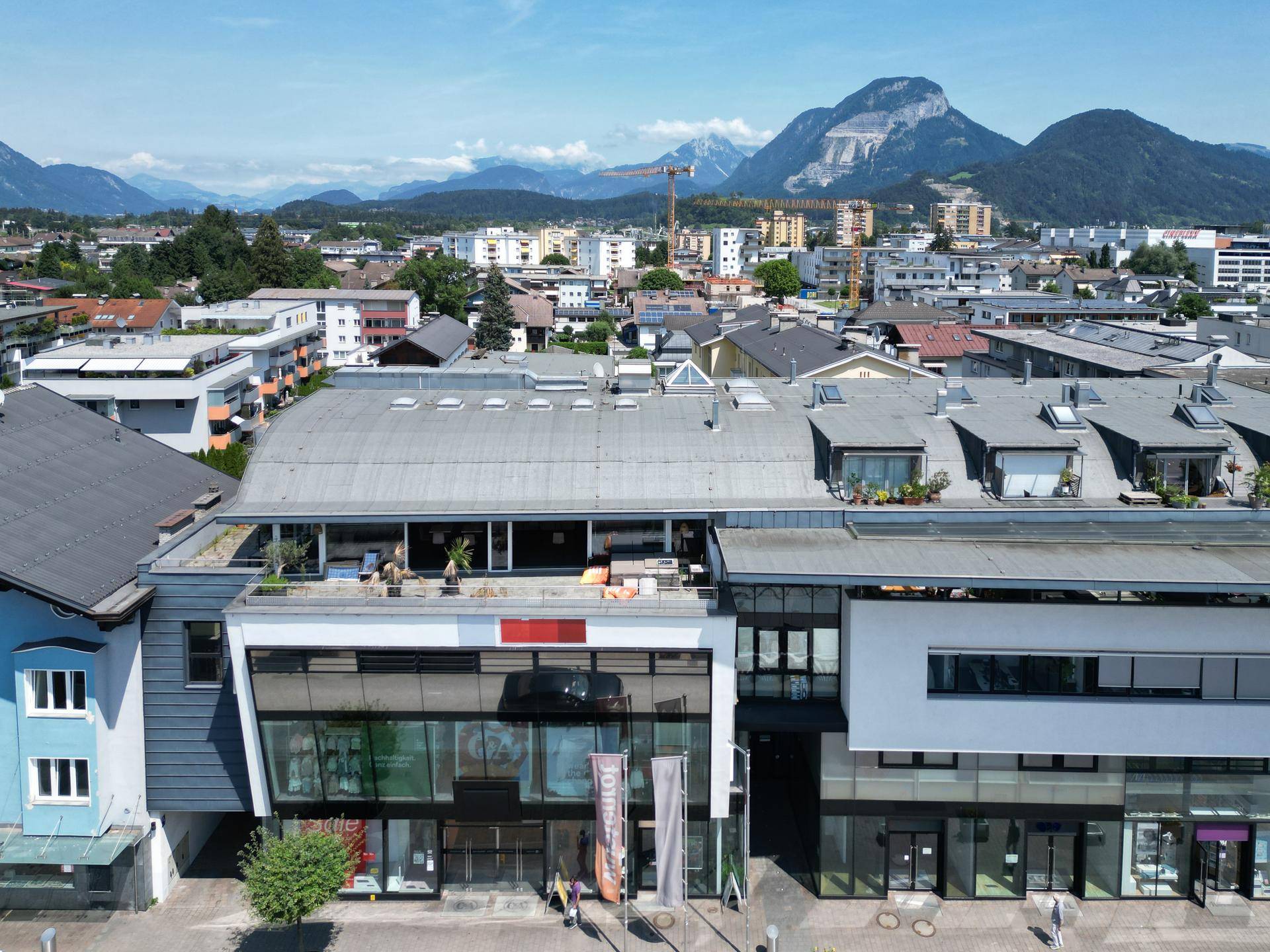 Luftaufnahme eines modernen Gebäudes mit Dachterrasse und Stadtblick vor einem weiten Bergpanorama.
