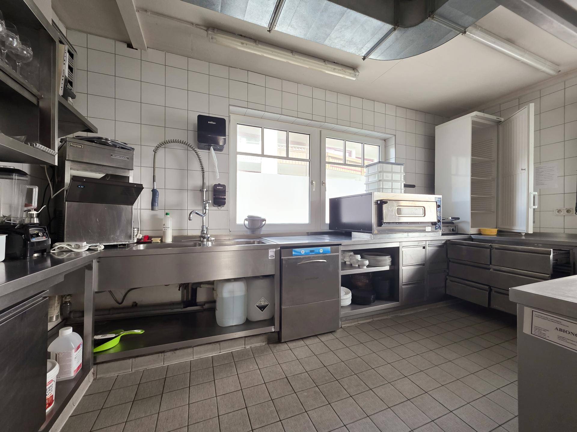 Professionelle Küche mit Edelstahlgeräten, Spülmaschine, Backofen und gefliesten Wänden.