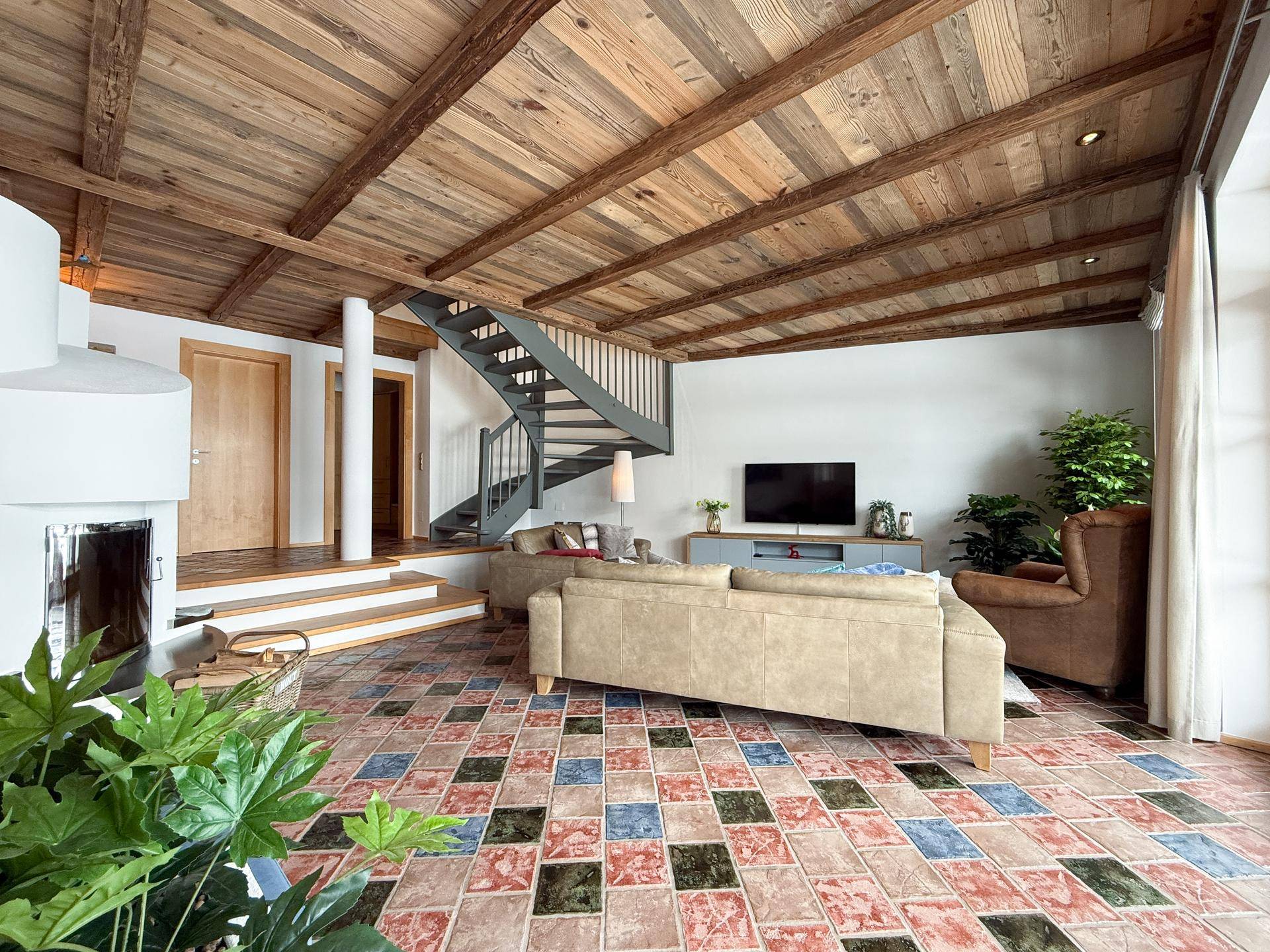 Geräumiges Wohnzimmer mit Holzbalkendecke, farbigen Fliesen und einem modernen Kaminofen.