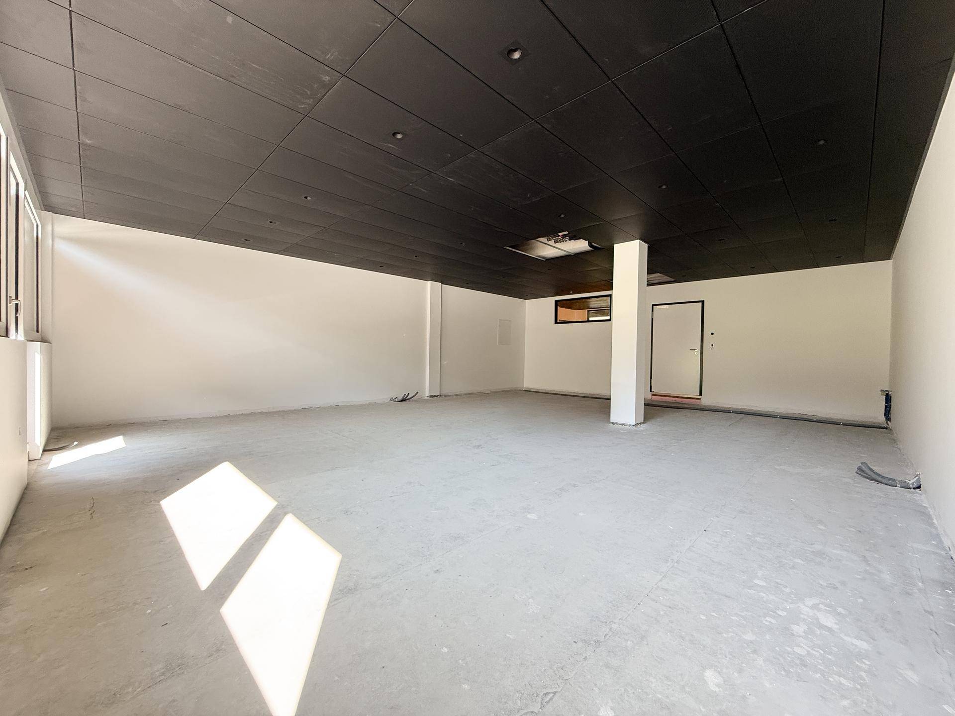 Heller, leerer Gewerberaum mit Betonboden und schwarzer Decke, durchflutet von natürlichem Tageslicht.