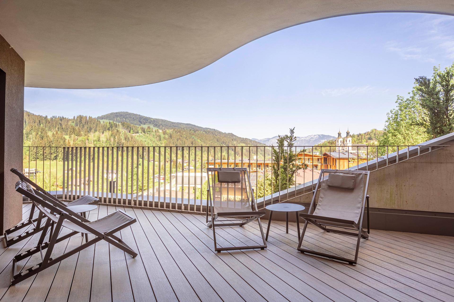Großer, geschwungener Balkon mit Holzdeck und Liegestühlen, bietet einen weiten Blick auf Berge und Wälder.