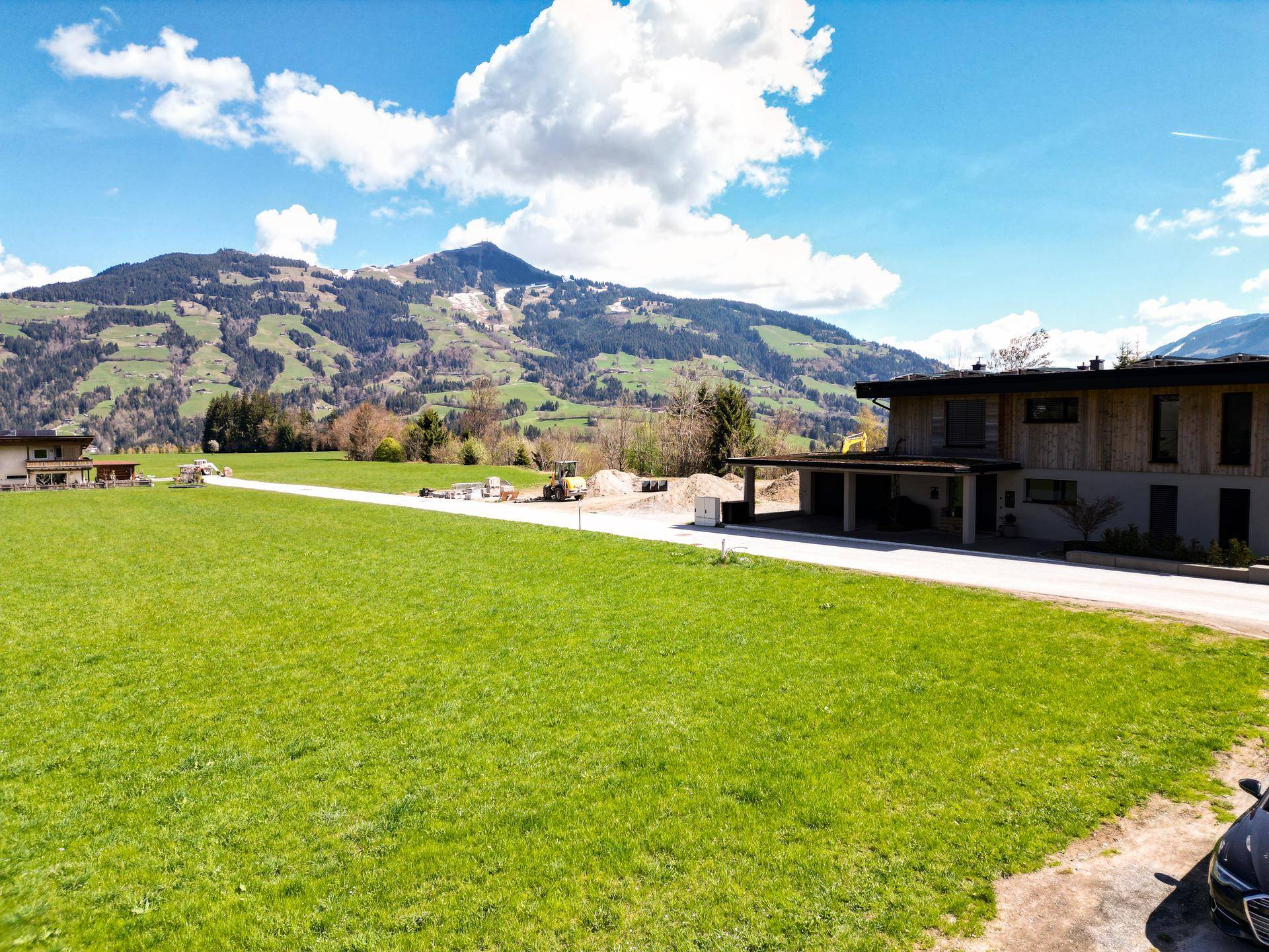 Modernes Wohnhaus mit Holzfassade und Carport, umgeben von grünen Wiesen und Bergen im Hintergrund.
