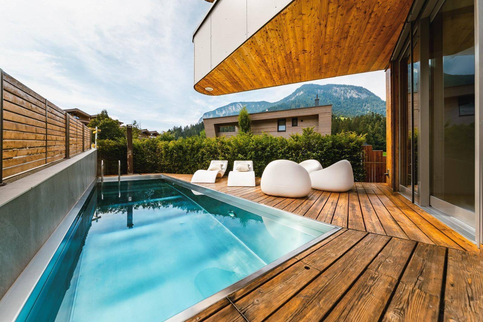 Außenpool mit Holzdeck und Liegen, umgeben von grüner Landschaft und Bergen im Hintergrund.