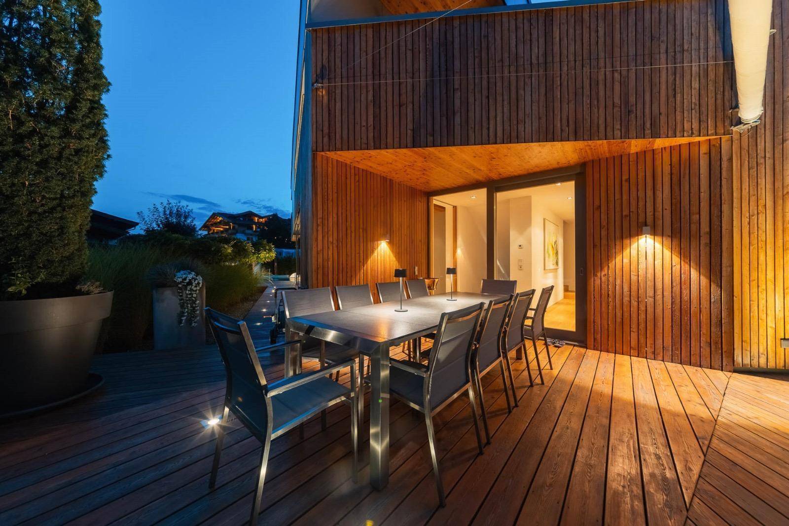 Beleuchtete Holzterrasse am Abend mit großem Esstisch und Zugang zum Innenbereich.
