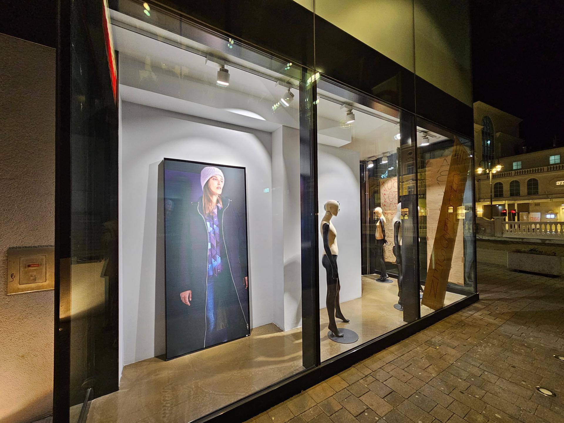 Nahaufnahme eines beleuchteten Schaufensters mit Schaufensterpuppen und einer digitalen Anzeige, sichtbar von der Straße.