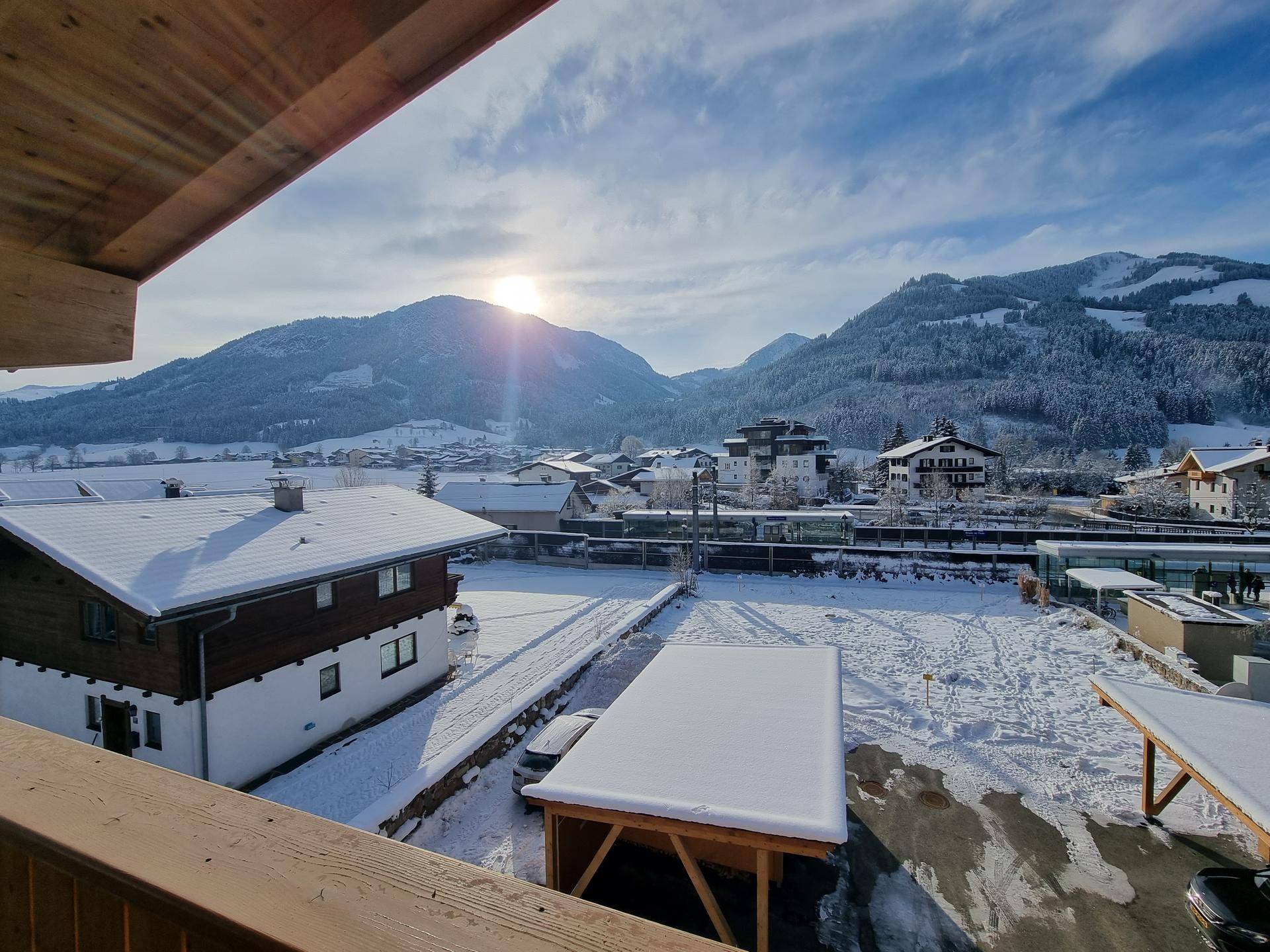 Sonniger Ausblick vom Balkon auf eine verschneite Berglandschaft und umliegende Häuser.