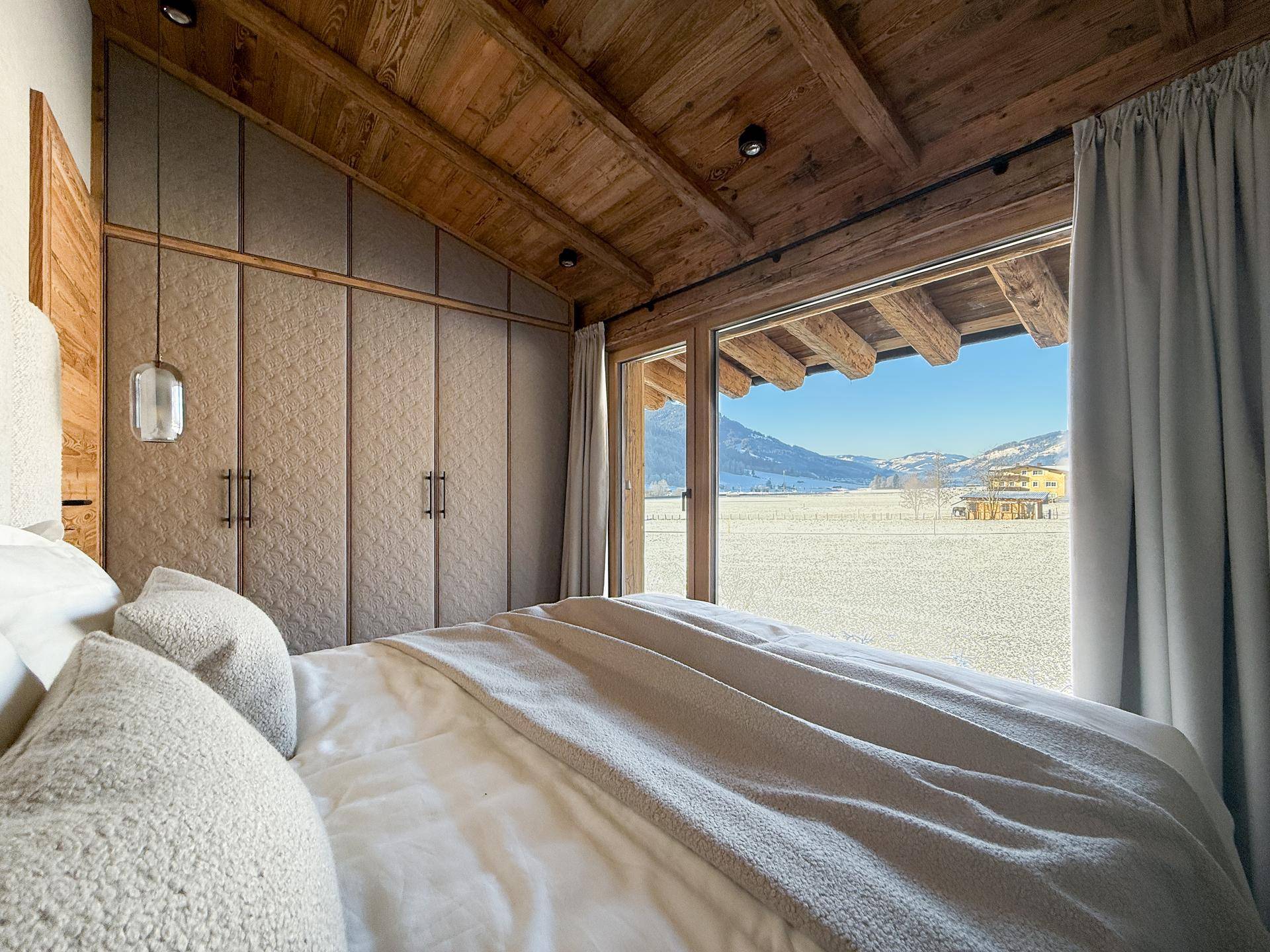 Schlafzimmer mit Einbauschränken, Holzbalkendecke und weitem Blick auf die verschneite Berglandschaft.