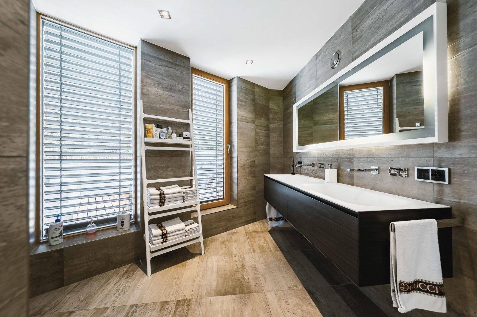 Modernes Badezimmer mit Doppelwaschbecken, großen Fliesen und zwei Fenstern für Tageslicht.