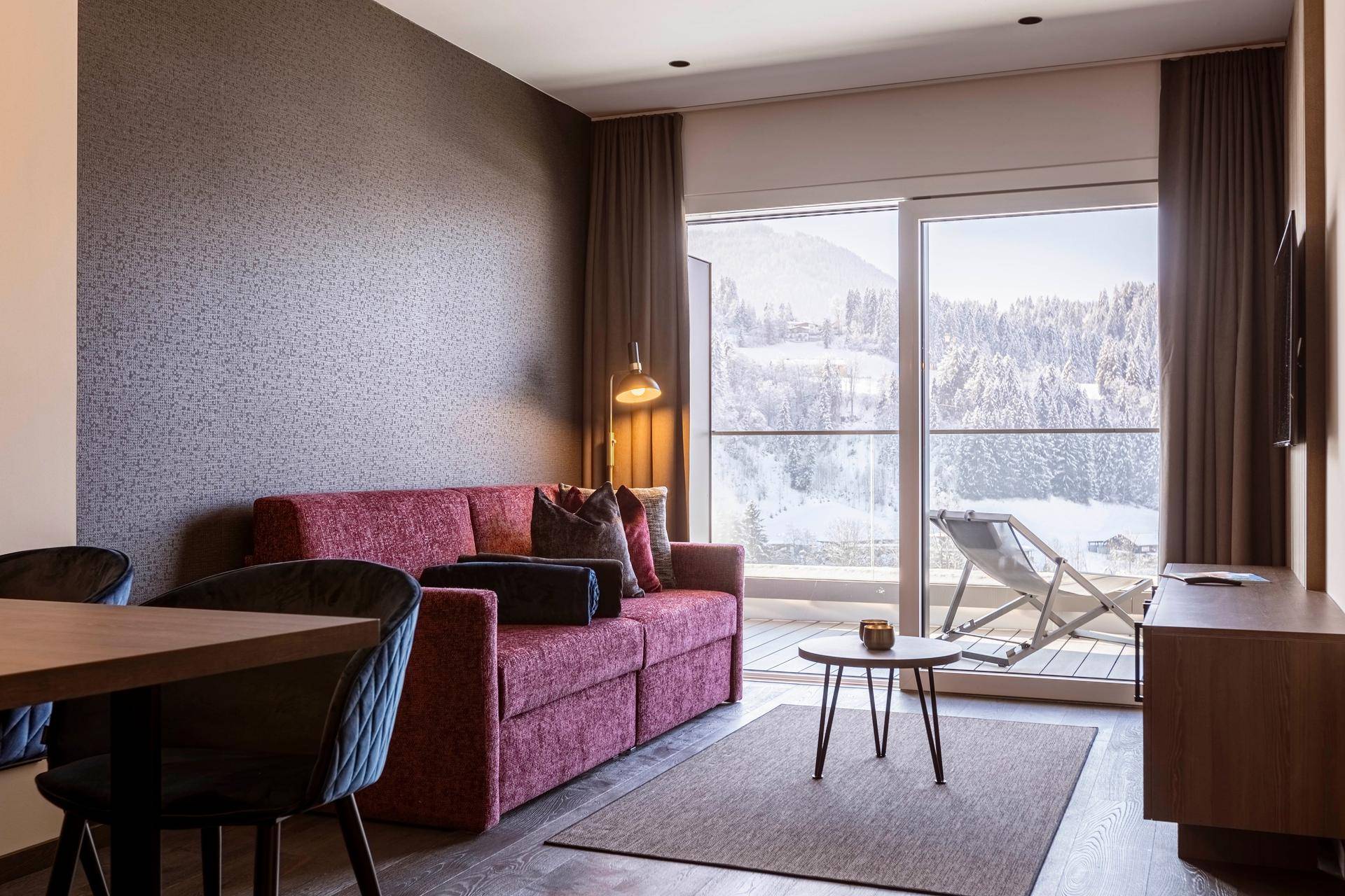 Helles Wohnzimmer mit Sofa, Balkonzugang und Liegestuhl mit Bergblick.