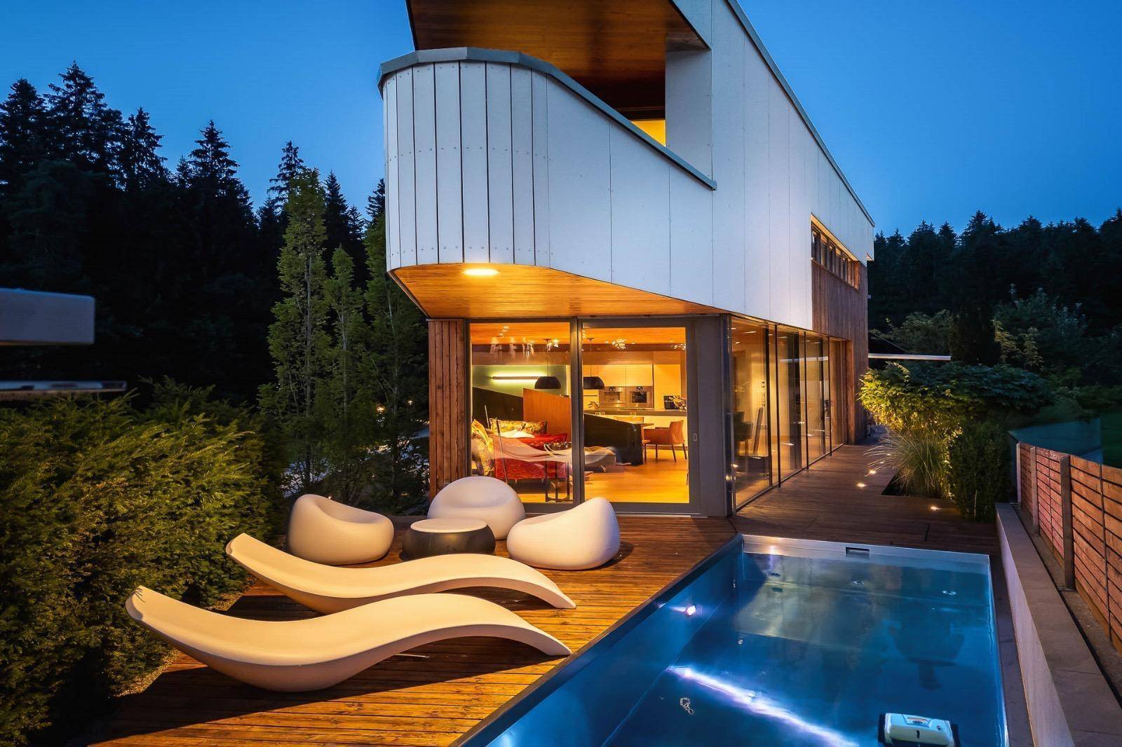 Moderner Außenpool mit Holzdeck und Liegen, angrenzend an die Glasfassade des Hauses.