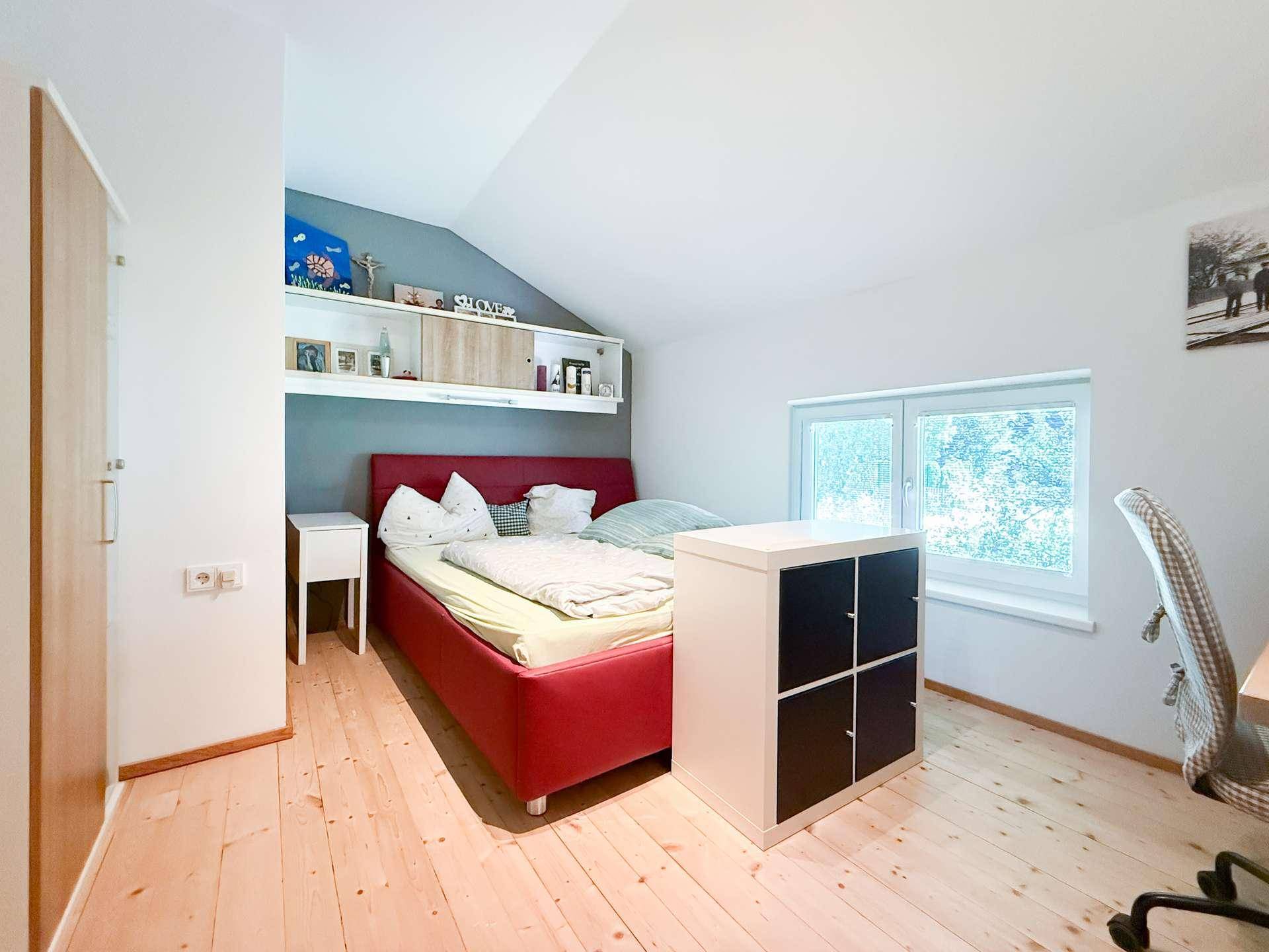 Schlafzimmer im Dachgeschoss mit rotem Bett, Stauraum und Fenster mit Grünblick.