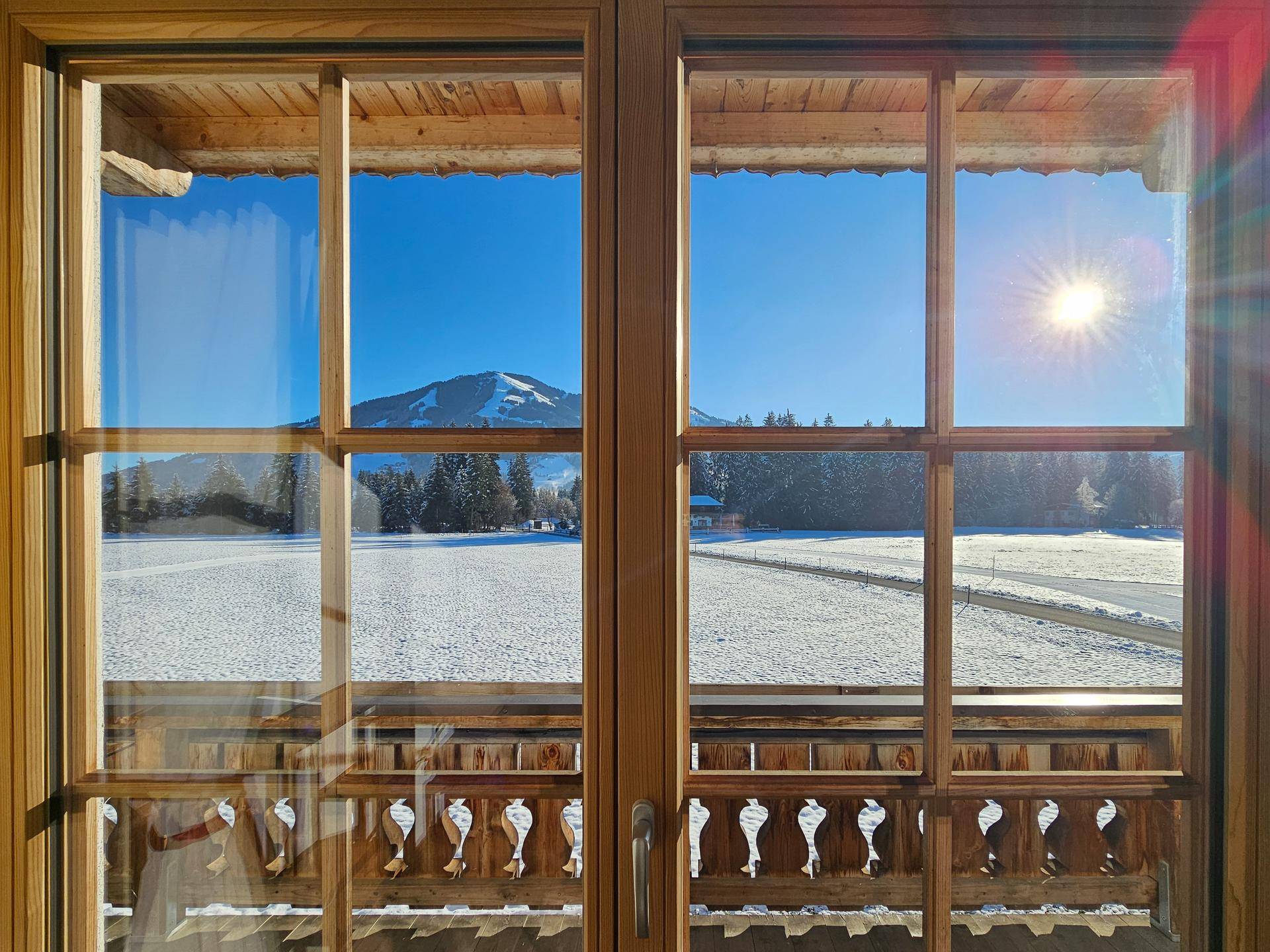Sonniger Ausblick durch ein Holzfenster auf eine weite, verschneite Landschaft mit Bergen unter blauem Himmel.