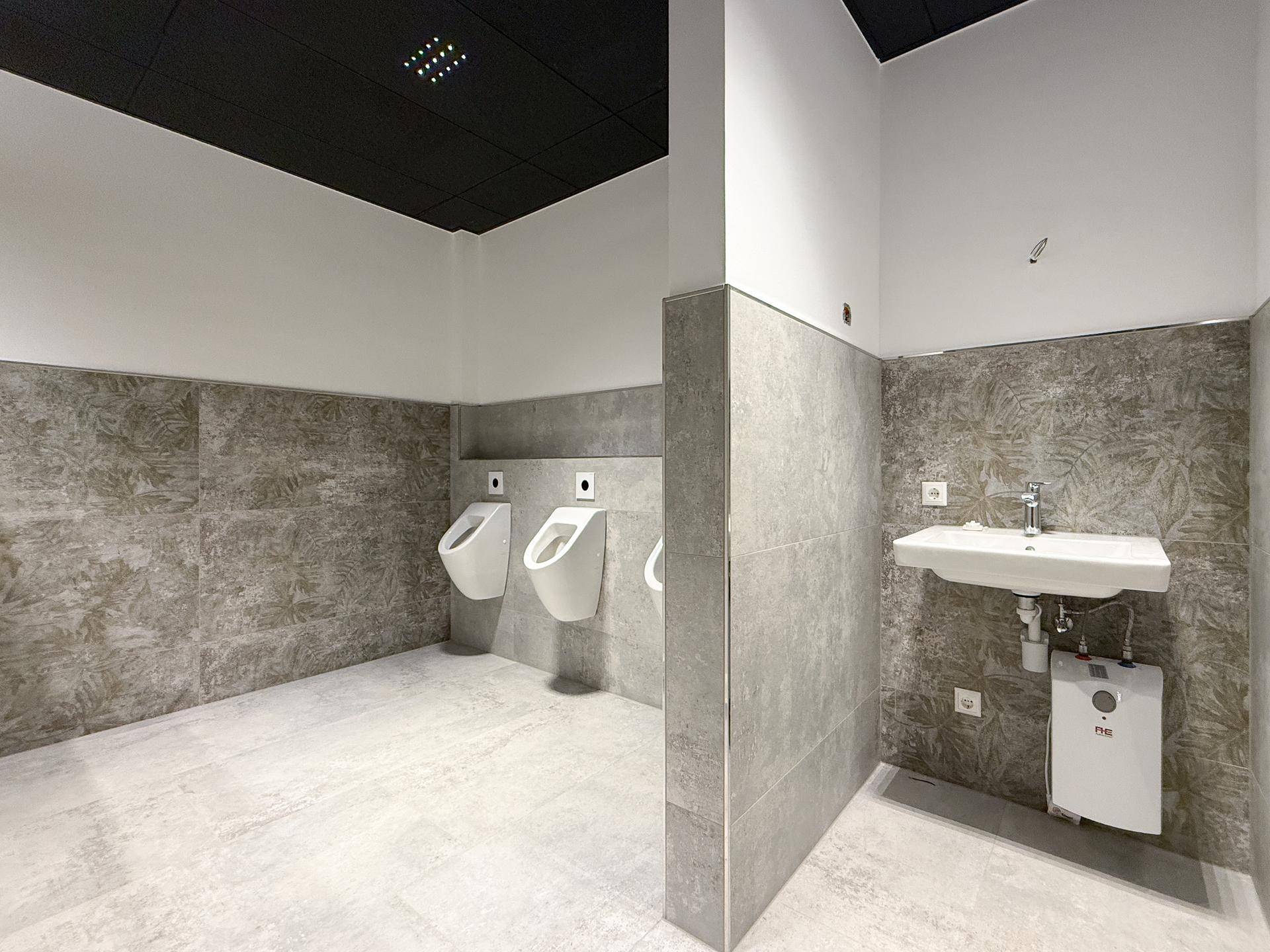 Moderner Herrenwaschraum mit Urinalen, Waschbecken und grauen Fliesen an Wänden und Boden.