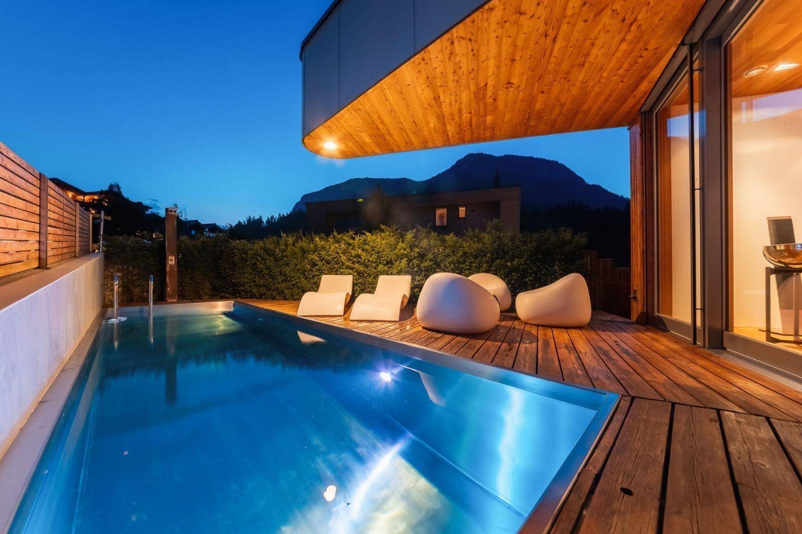 Außenpool mit Holzdeck und modernen Liegen, umgeben von grüner Landschaft und Bergen.