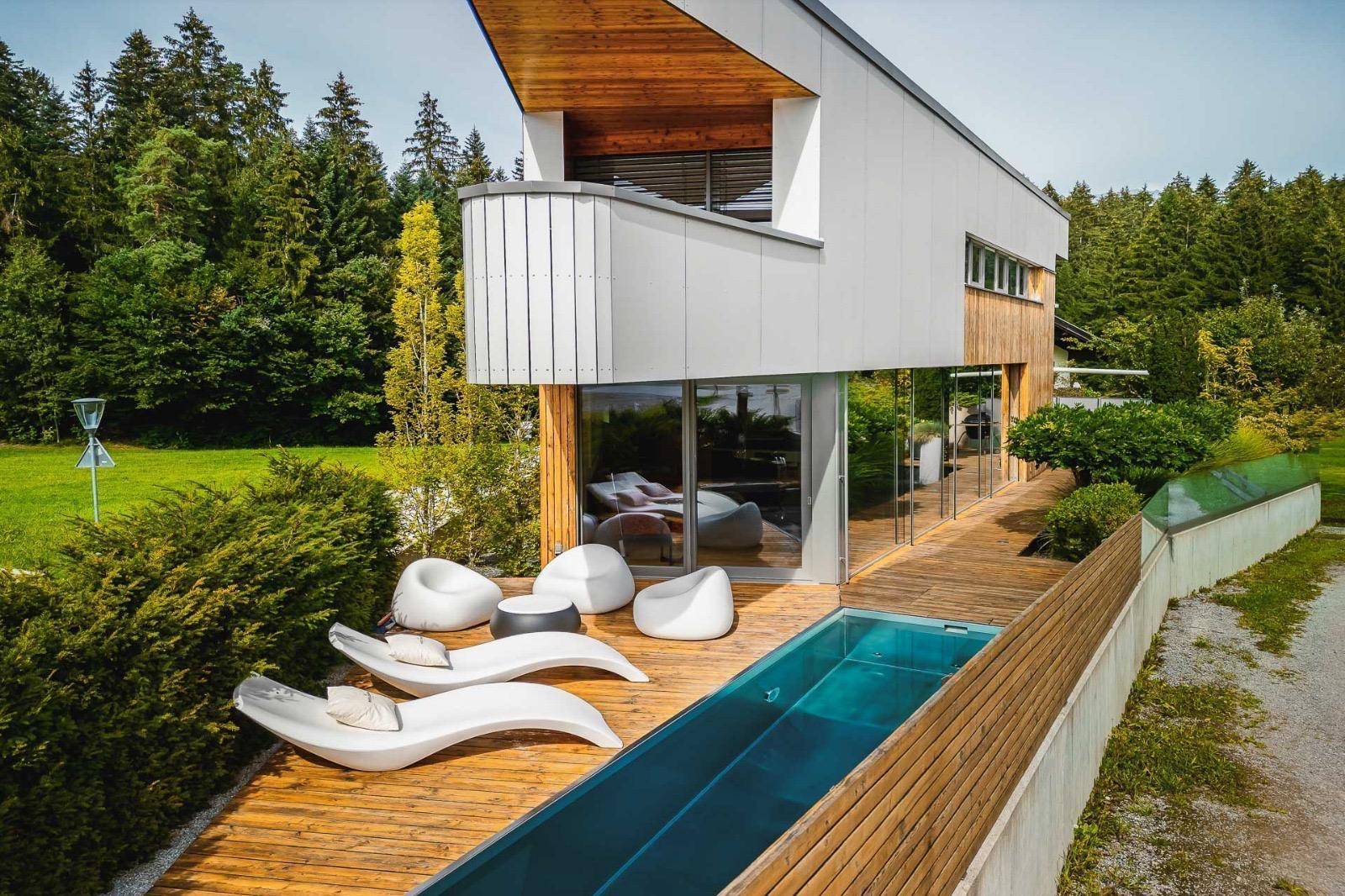 Außenpool mit Holzdeck und modernen Liegen, umgeben von grüner Natur unter blauem Himmel.