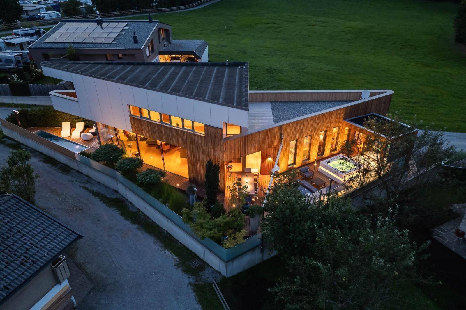 Luftaufnahme des modernen Hauses mit Holzfassade, Pool, Whirlpool und umliegender Natur bei Dämmerung.