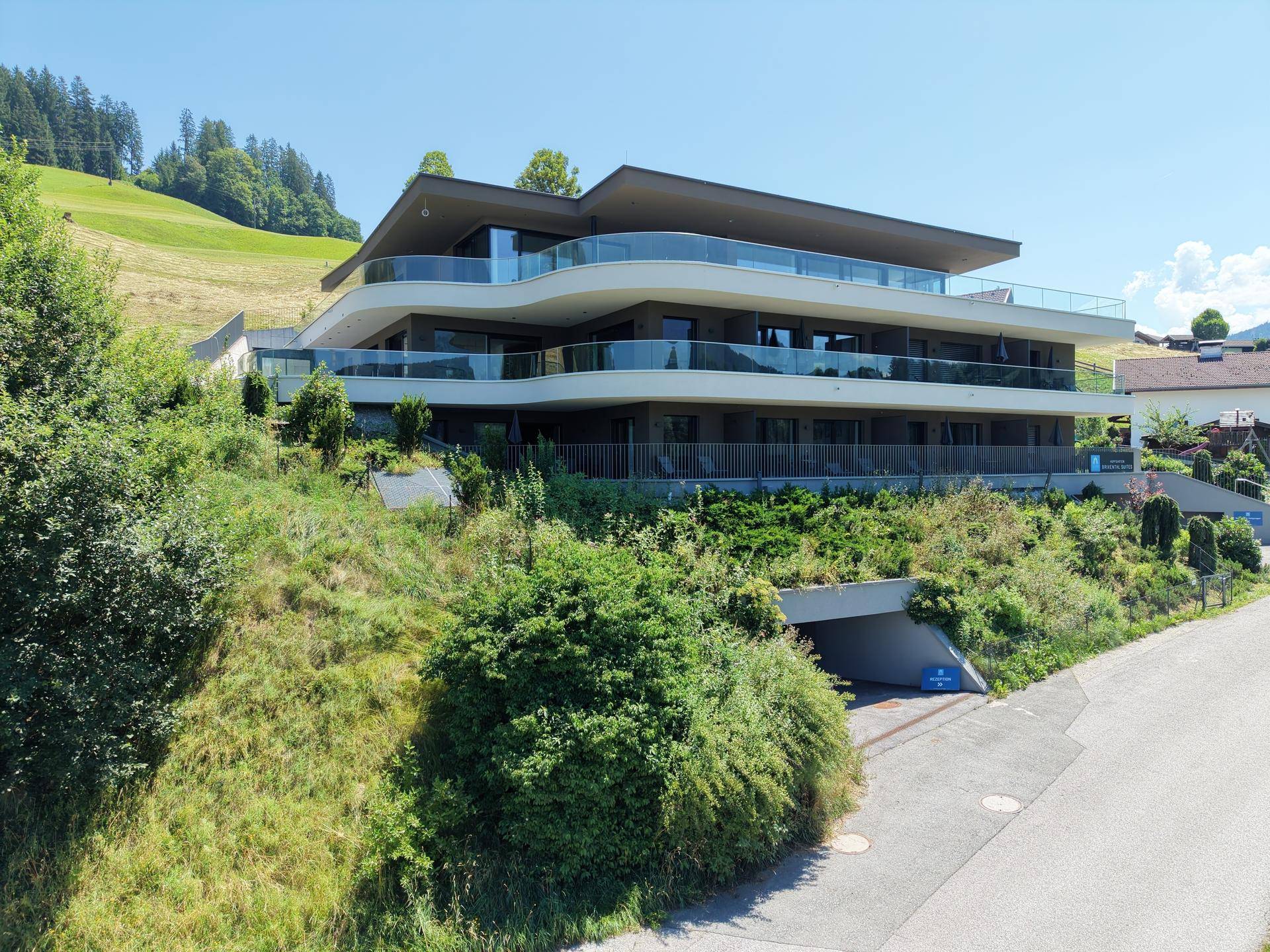 Modernes Apartmentgebäude mit geschwungenen Balkonen und viel Grün in alpiner Umgebung.