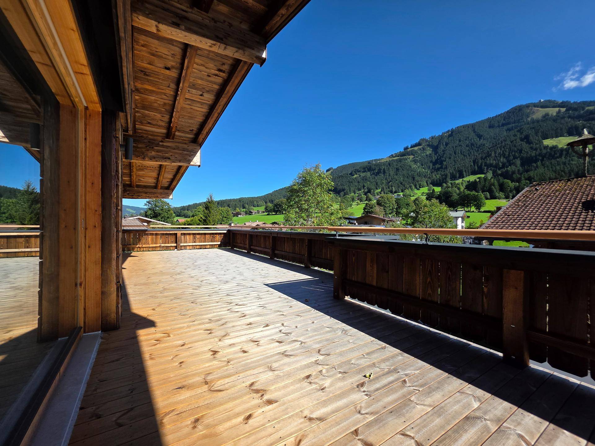 Geräumige Holzterrasse mit Panoramablick auf die Alpen und die grüne Natur unter strahlendem Sonnenschein.