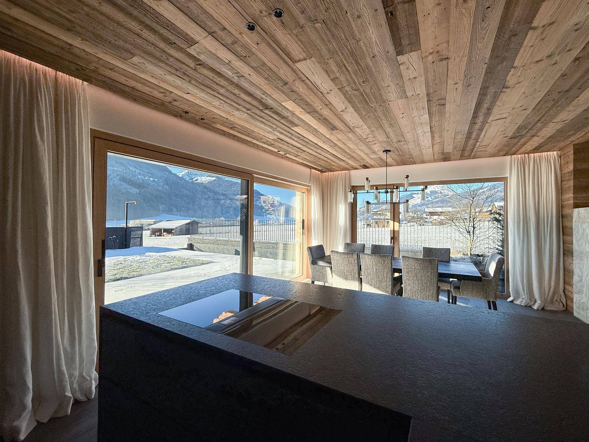 Moderne Küche mit Kochinsel und Essbereich, umgeben von großen Fenstern mit Winterlandschaftsblick.