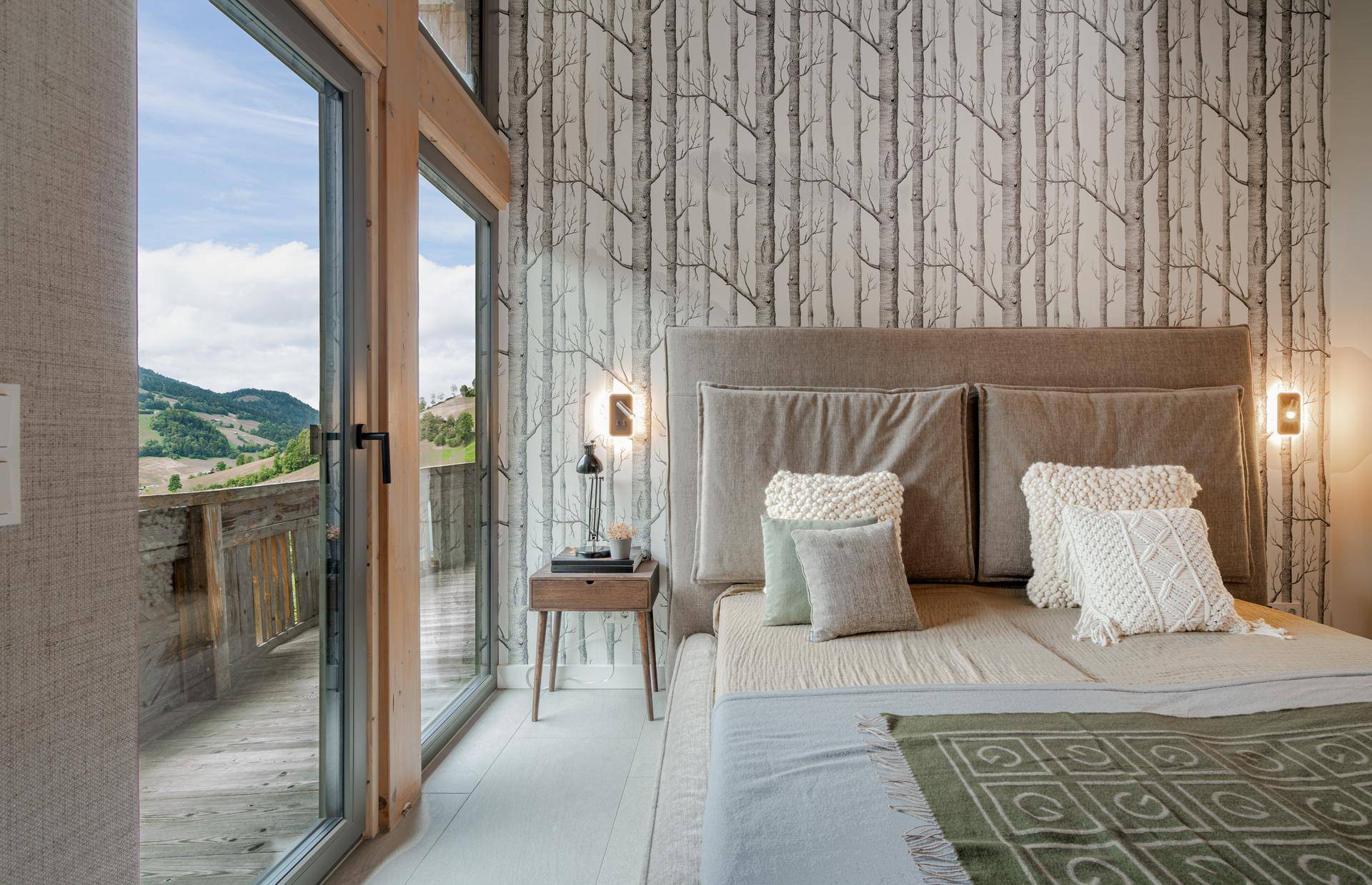 Schlafzimmer mit Tapete im Birkenmuster, großem Bett und raumhohen Fenstern mit Bergblick.