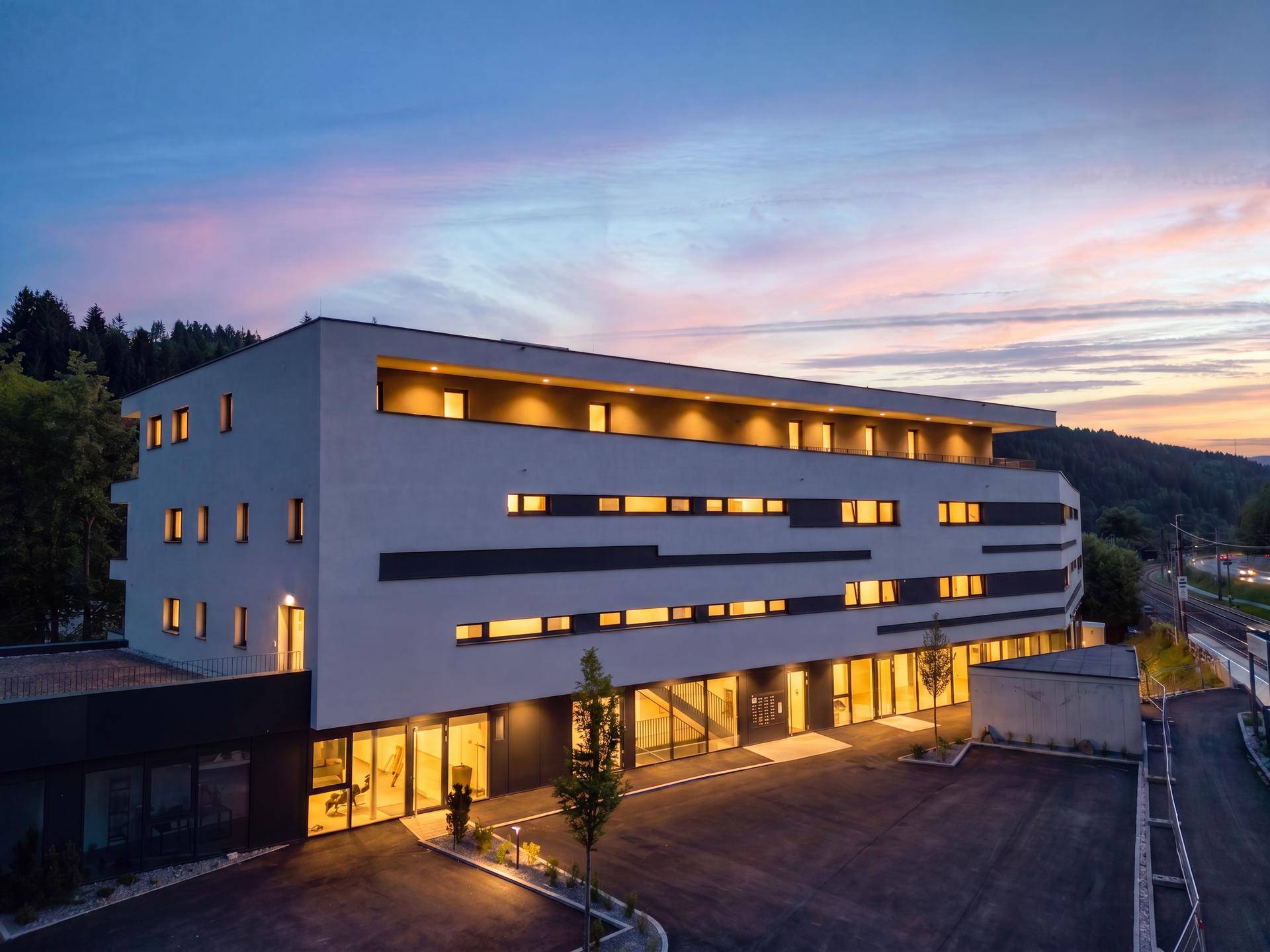 Modernes Gebäude mit beleuchteter Fassade bei Sonnenuntergang, umgeben von Hügeln.