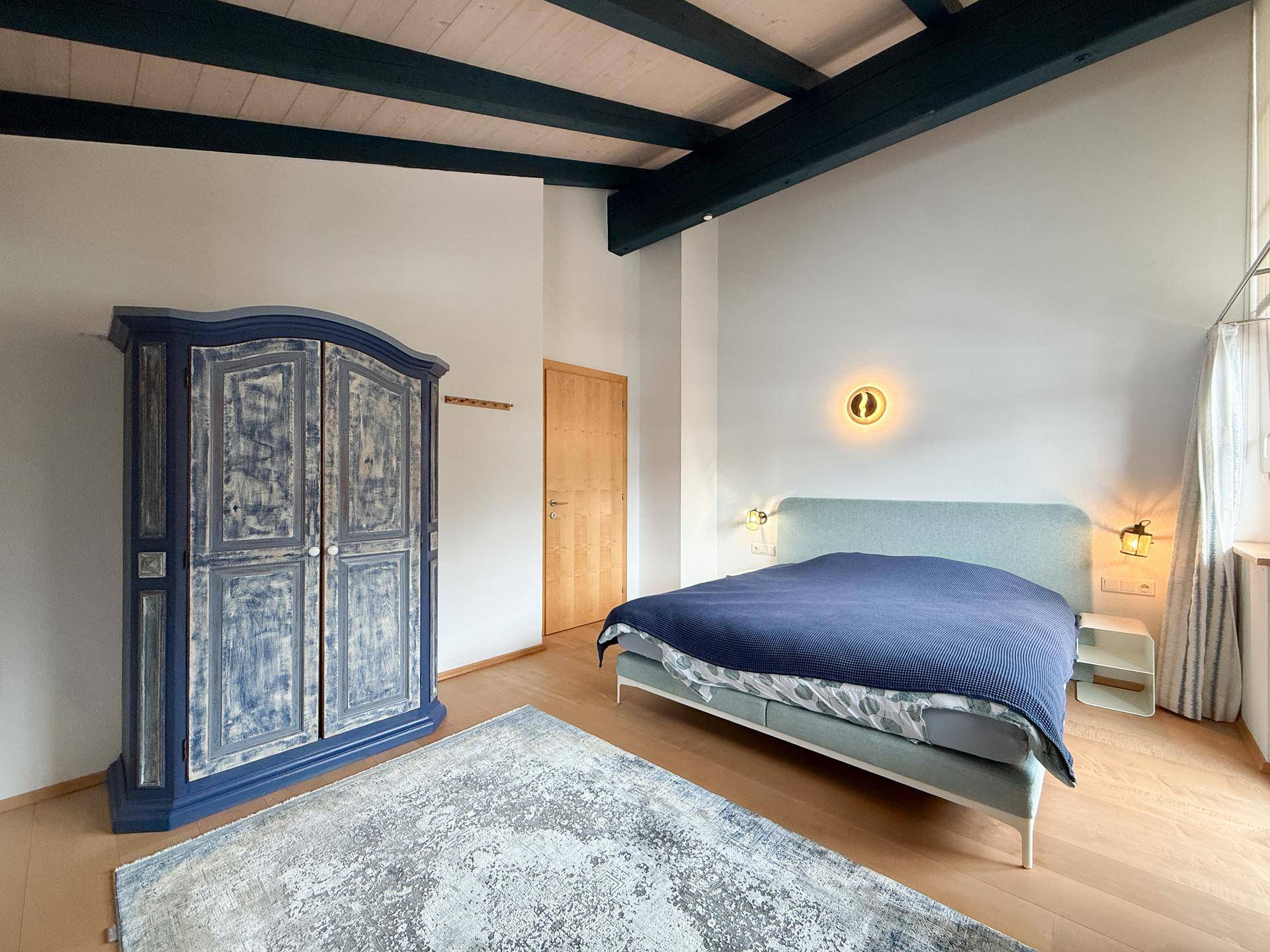 Schlafzimmer mit Holzbalkendecke, blauem Kleiderschrank und hellem Holzboden.
