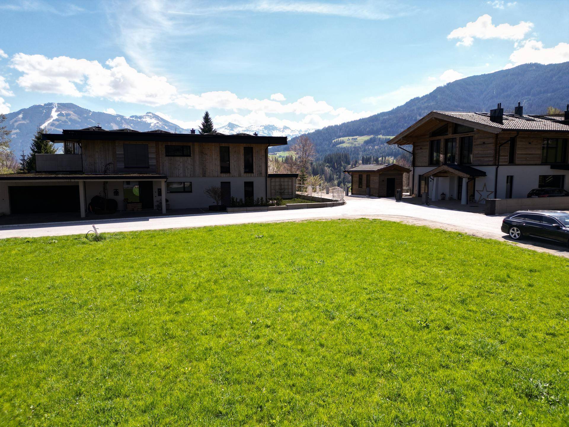 Zwei moderne Wohnhäuser mit Holzfassaden und Carports, eingebettet in eine grüne Landschaft mit Bergblick.