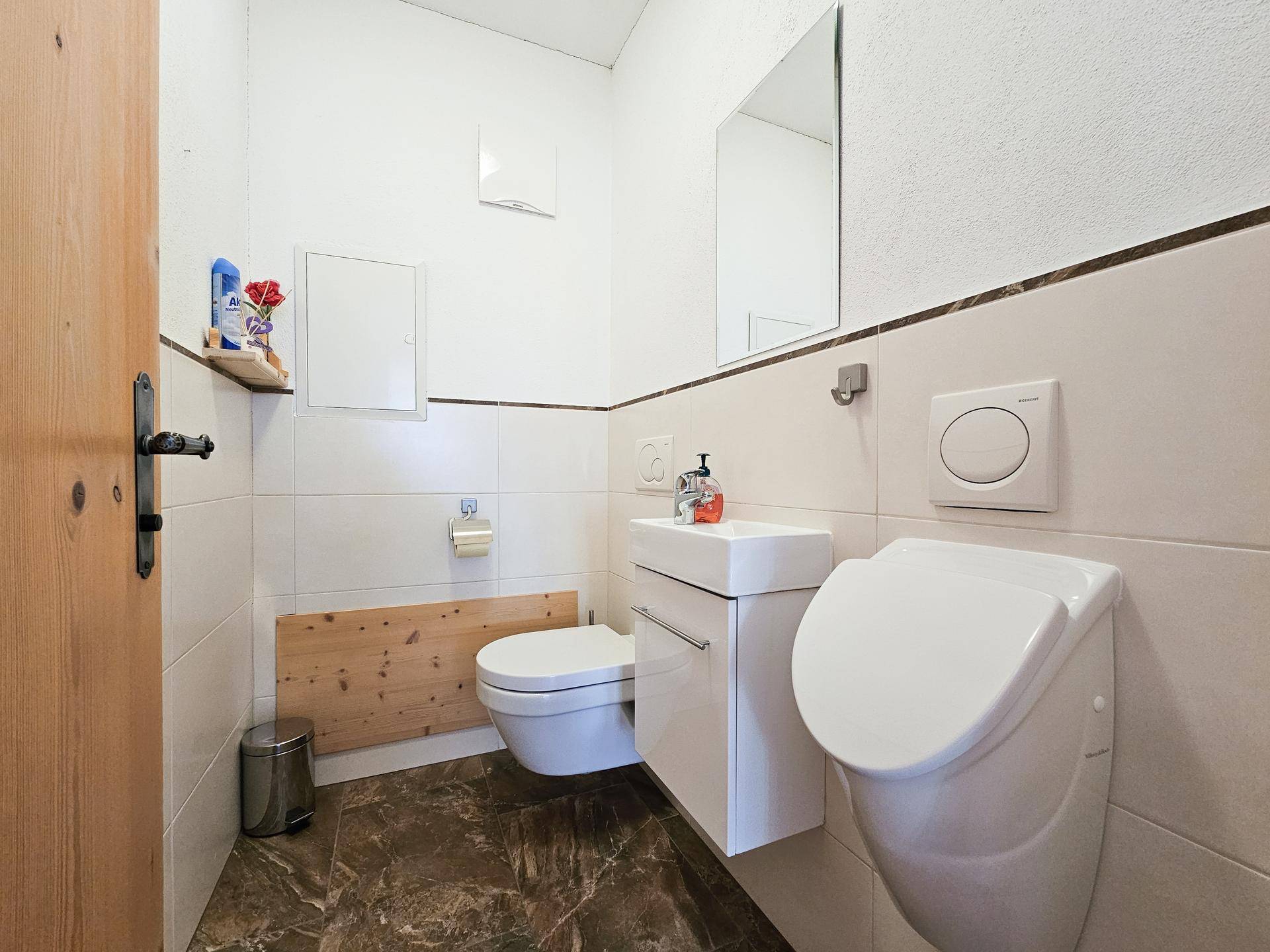 Kleines Gäste-WC mit Toilette, Urinal und Handwaschbecken, in hellen und dunklen Fliesen gehalten.