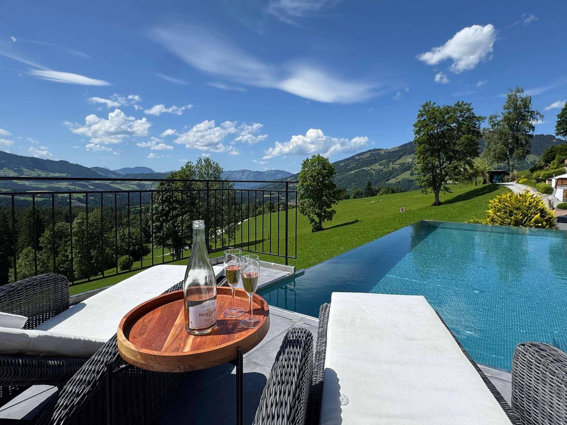 Infinity-Pool mit Liegestühlen und atemberaubendem Blick auf die umliegende Berglandschaft unter blauem Himmel.