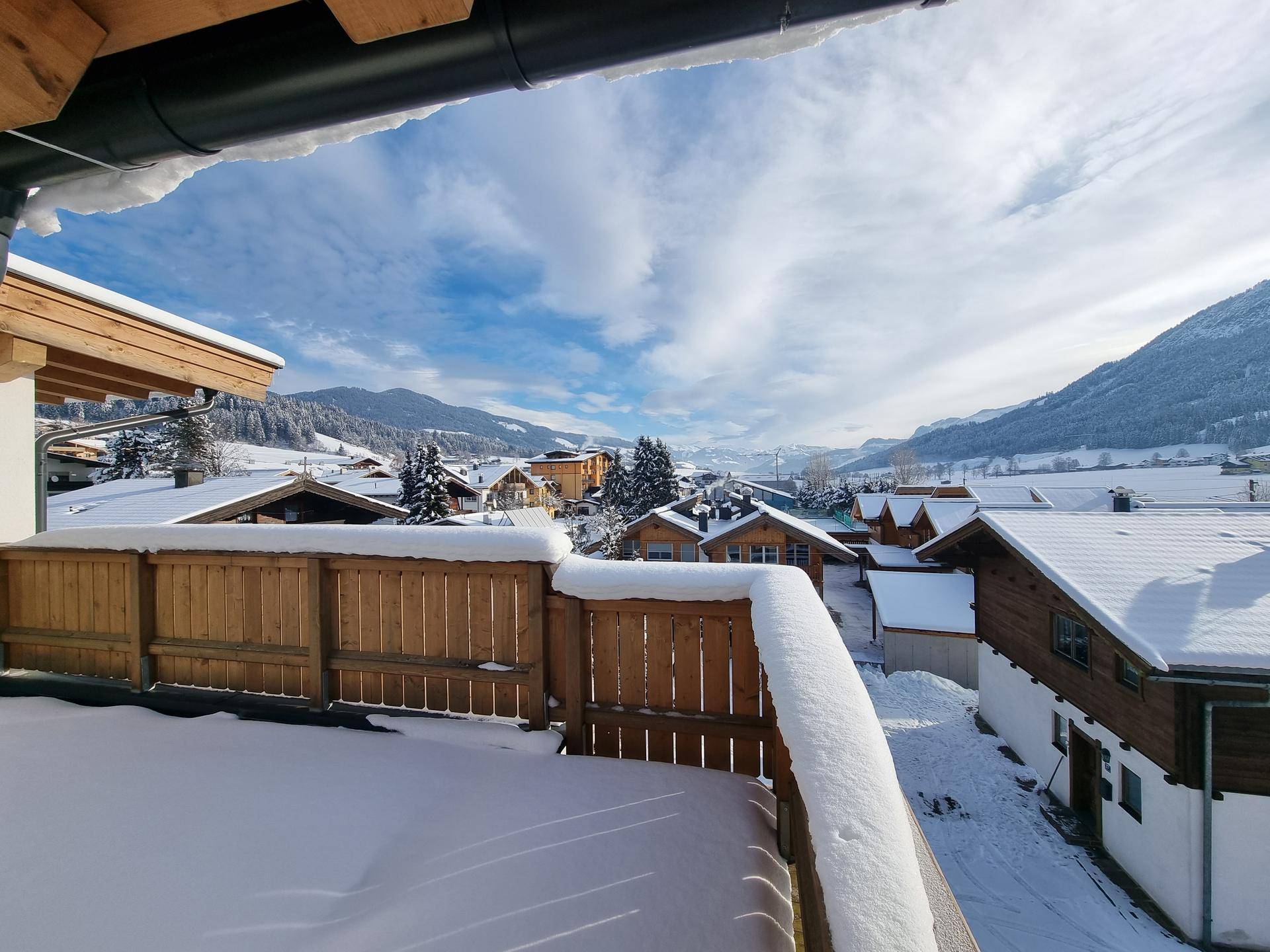 Verschneiter Holzbalkon mit weitem Blick über das winterliche Dorf und die umliegenden Berge.