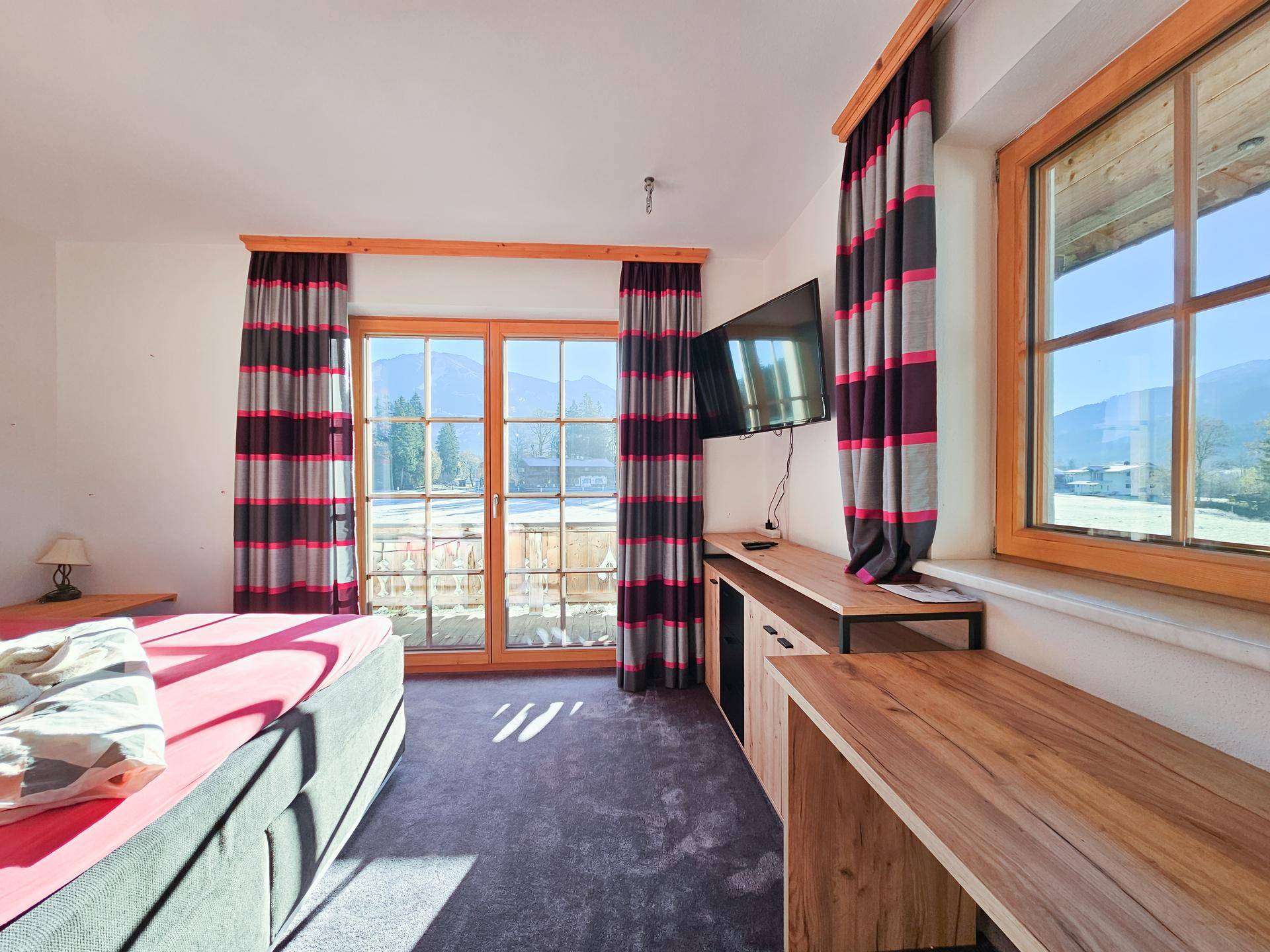Schlafzimmer mit Balkonzugang, Fernseher und einem Schreibtischbereich, der viel Tageslicht erhält.