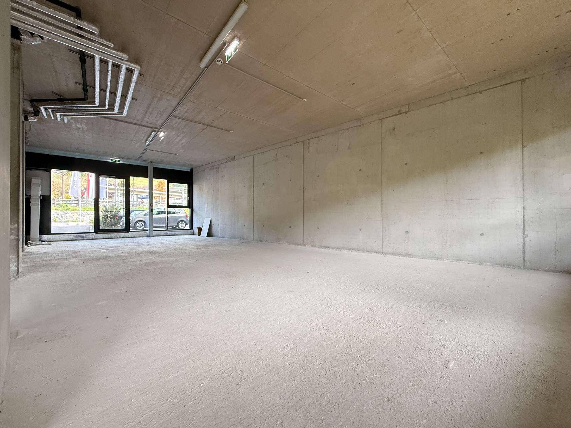 Leerer Innenraum im Rohbau mit Sichtbeton, freiliegenden Leitungen und großen Glasfronten zur Straße mit einem Auto.