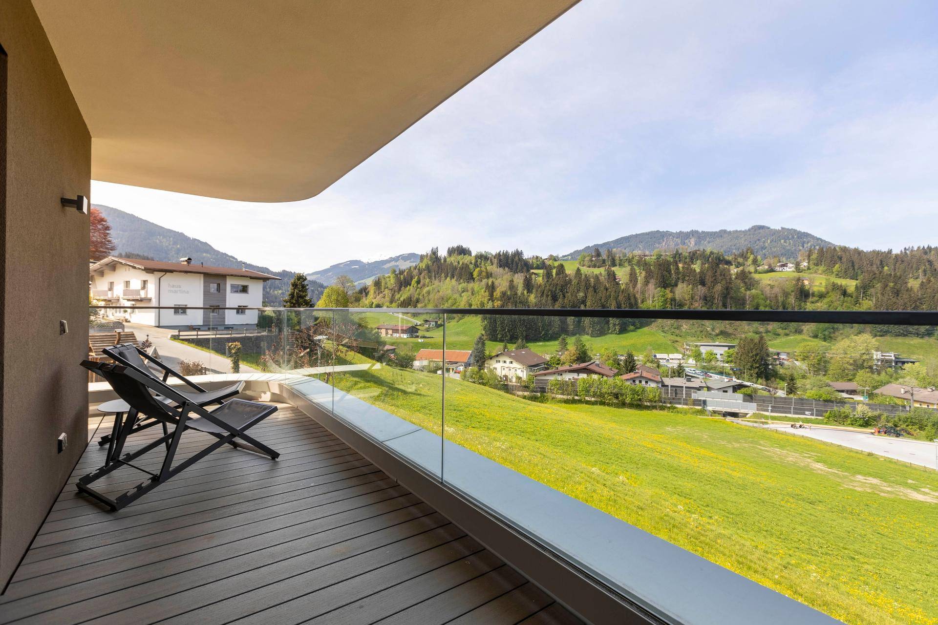 Balkon mit Glasgeländer und Liegestühlen, bietet einen weiten Panoramablick über grüne Hügel und Häuser.