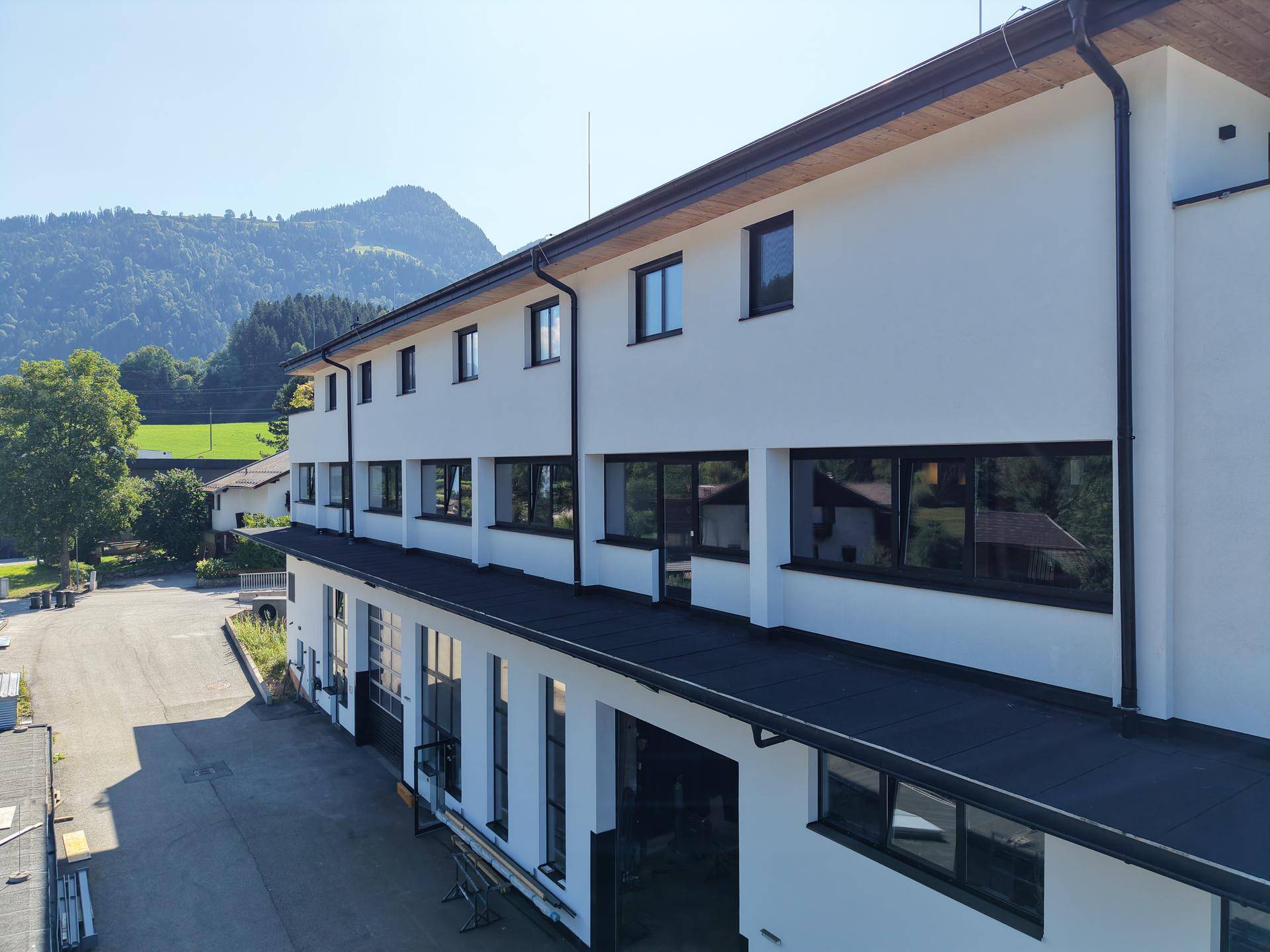 Seitenansicht des modernen Gebäudes mit mehreren Etagen, weißen Fassaden und dunklen Fensterrahmen vor Bergkulisse.