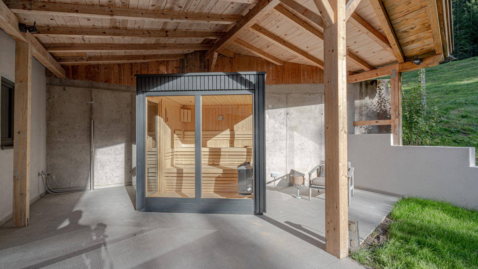 Externe Sauna mit Glasfront in einem überdachten Außenbereich, umgeben von Natur.
