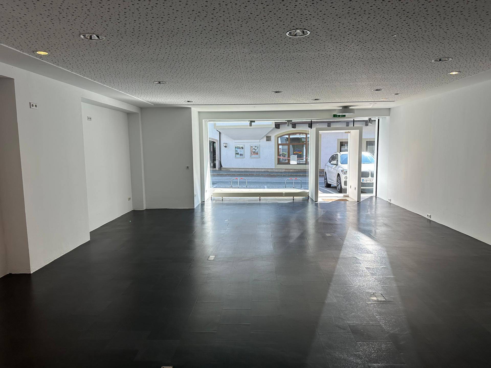 Offener Gewerberaum mit dunklem Boden, weißen Wänden und breiter Glasfront zur belebten Straße.