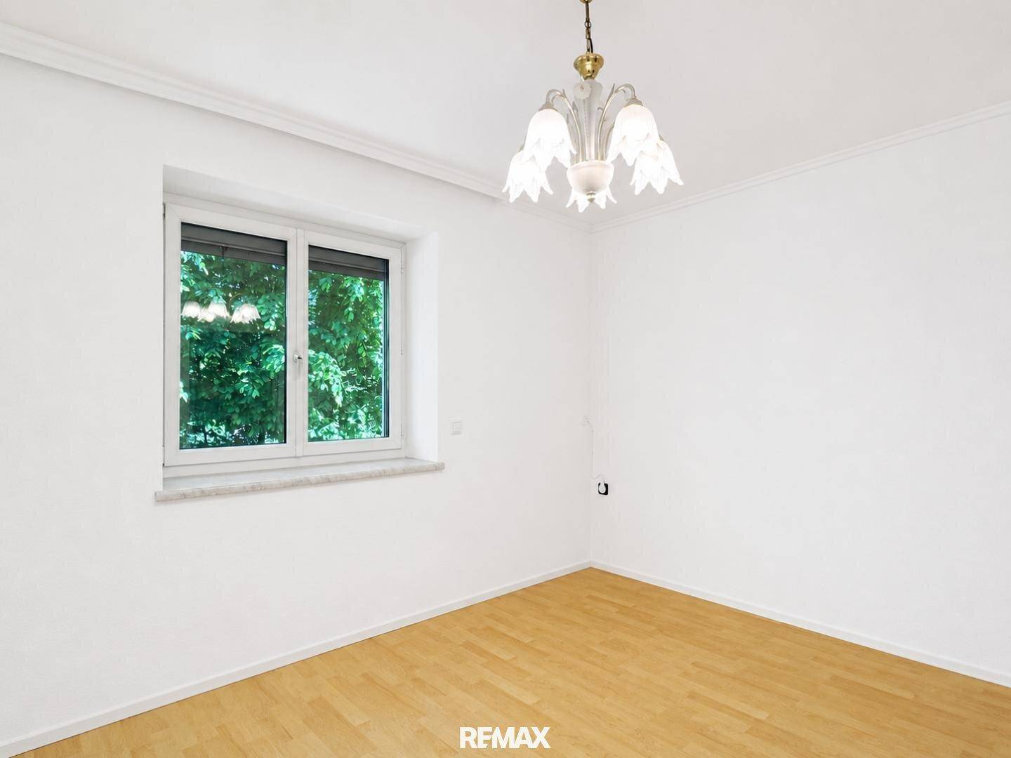 Helles Zimmer mit weißen Wänden, Holzfußboden und Blick ins Grüne durch das Fenster.