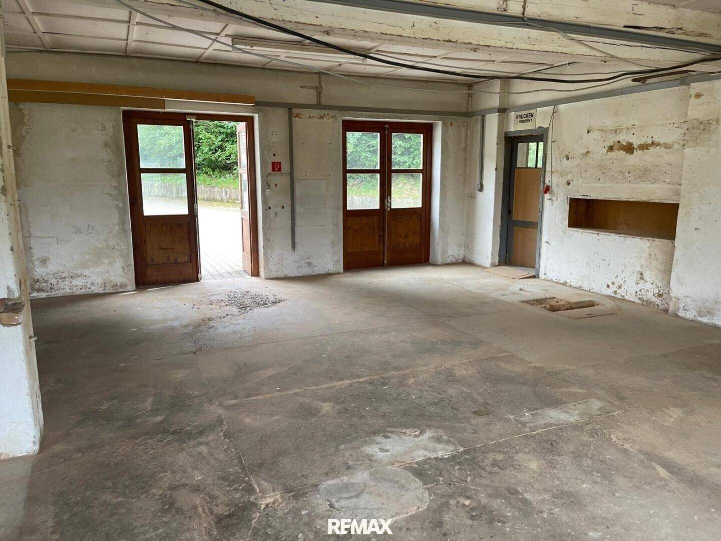 Großer, leerer Innenraum mit Betonboden und mehreren Holztüren, der Renovierungsbedarf zeigt.