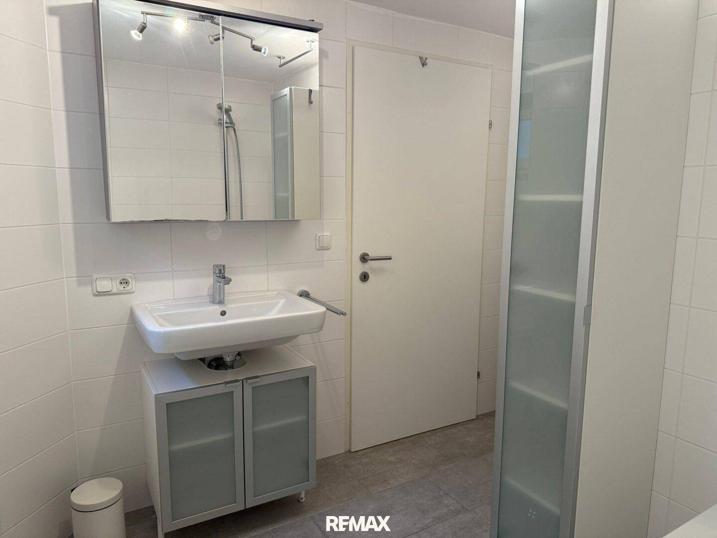 Helles Badezimmer mit weiß gefliesten Wänden, Einzelwaschbecken und Spiegelschrank.
