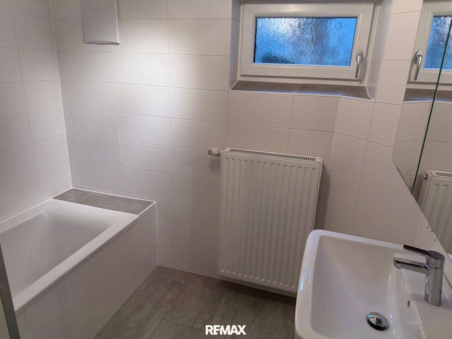 Badezimmer mit Badewanne, Fenster und Heizkörper, komplett gefliest in Weiß.
