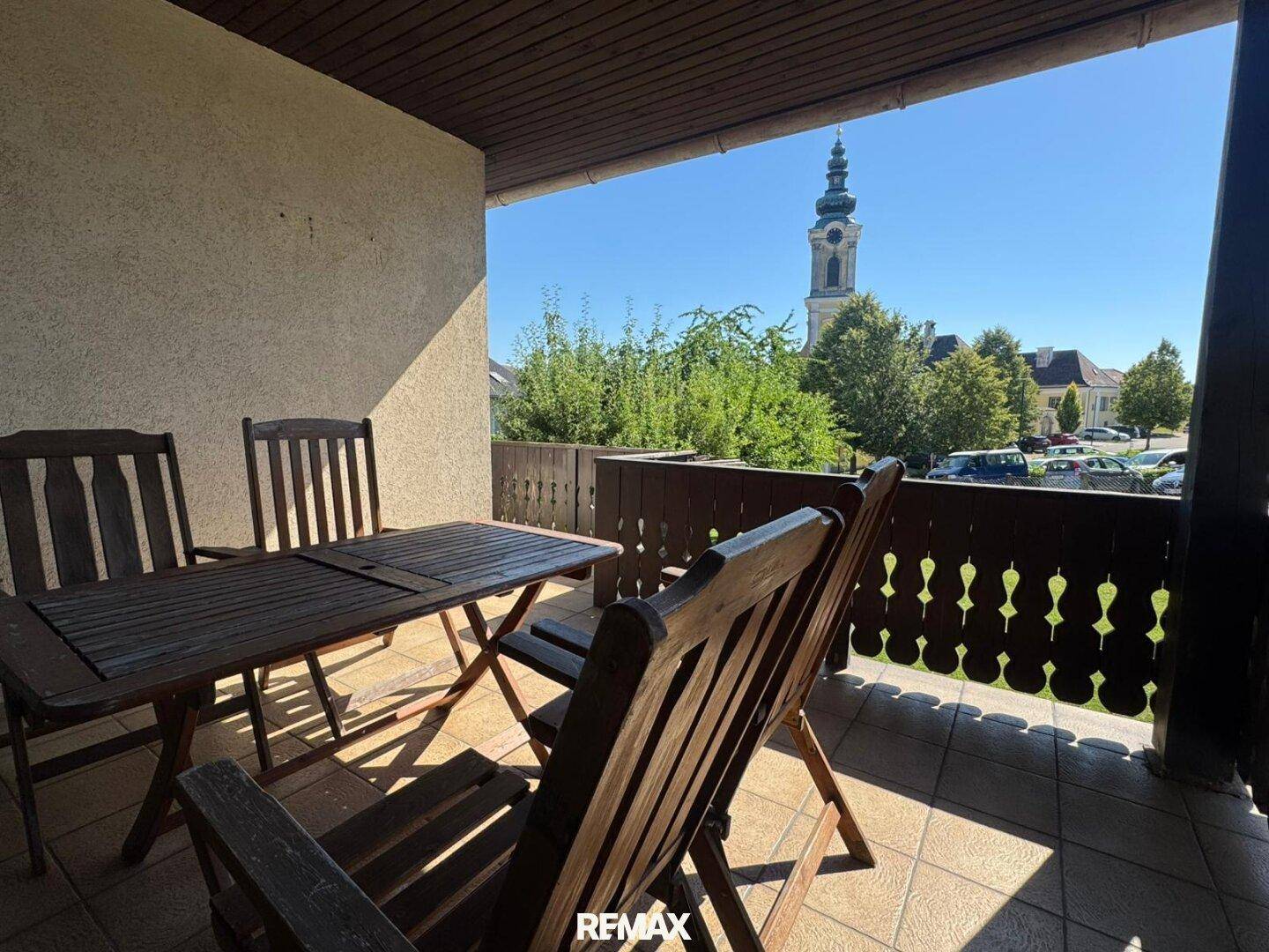 Gemütlicher Balkon mit Holztisch und Stühlen, bietet Blick auf die Umgebung und Kirchturm.