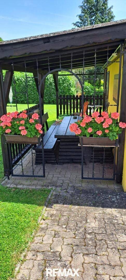 Überdachter Außenbereich mit Holztisch, Bänken und Blumenkästen unter einer Pergola im Garten.