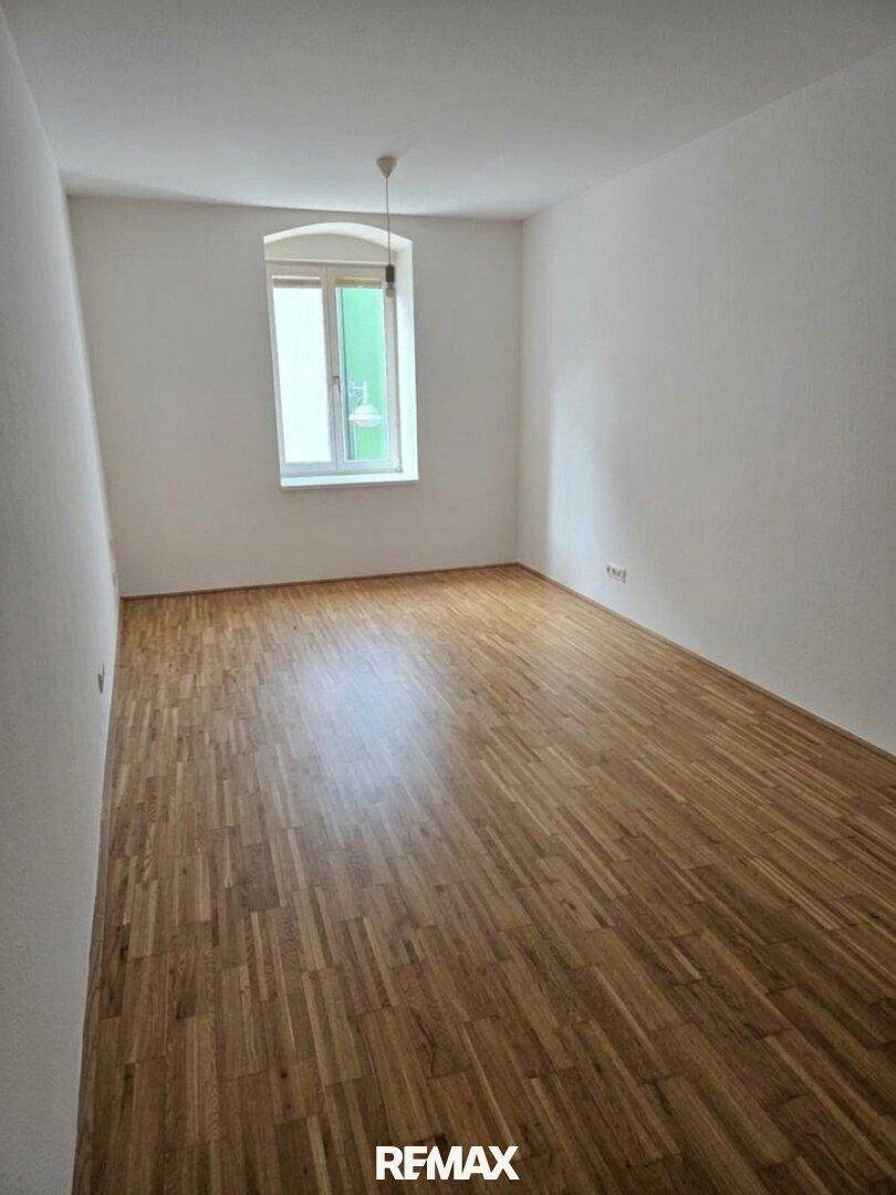 Leeres Zimmer mit hellem Parkettboden und einem Fenster, das viel Tageslicht hereinlässt.