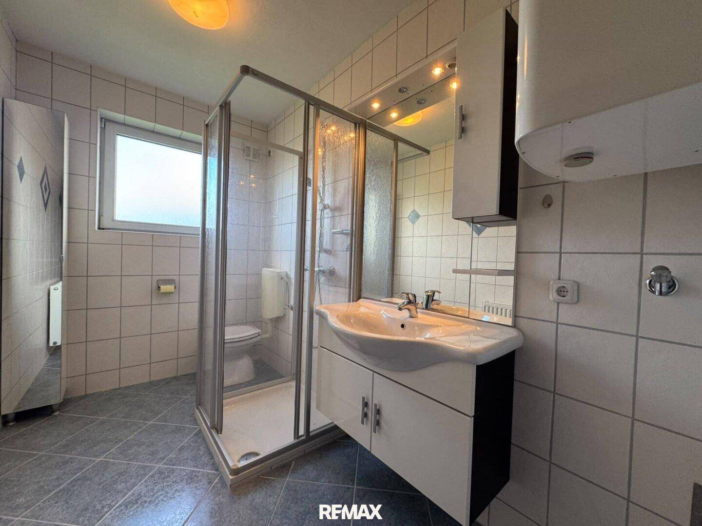 Badezimmer mit Duschkabine, Waschbecken und Fenster, gefliest in hellen Tönen.