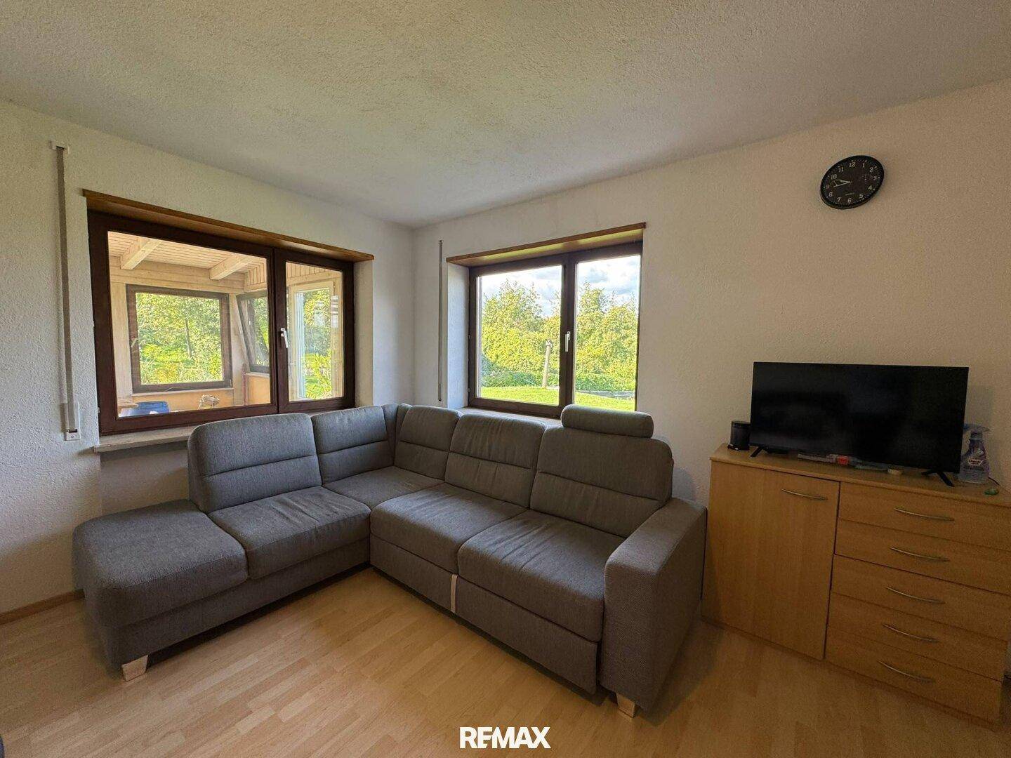 Gemütliches Wohnzimmer mit grauem Ecksofa, Holzboden und Blick ins Grüne.