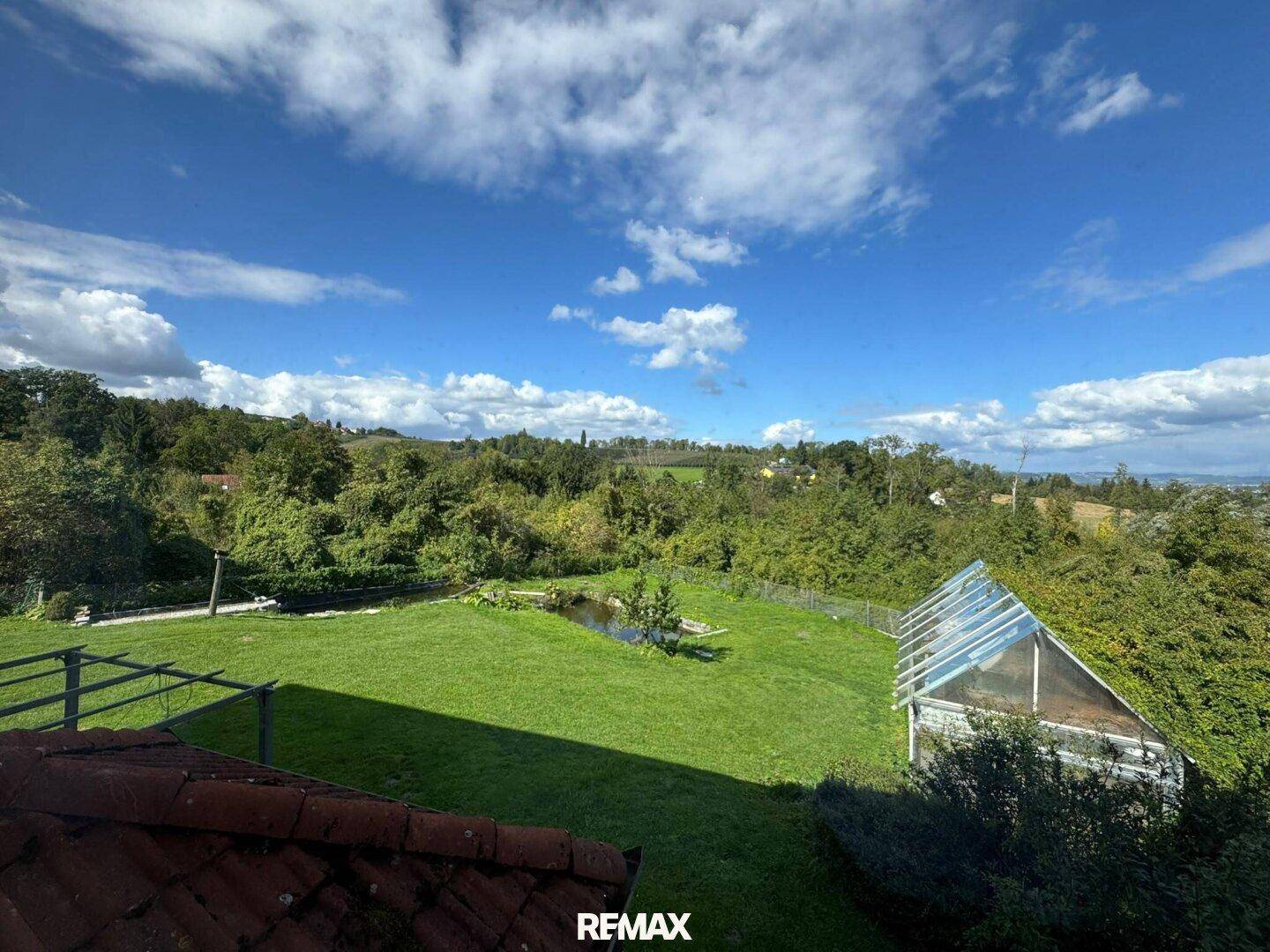 Panoramablick über den weitläufigen Garten mit Gewächshaus und Teich, umgeben von Natur.