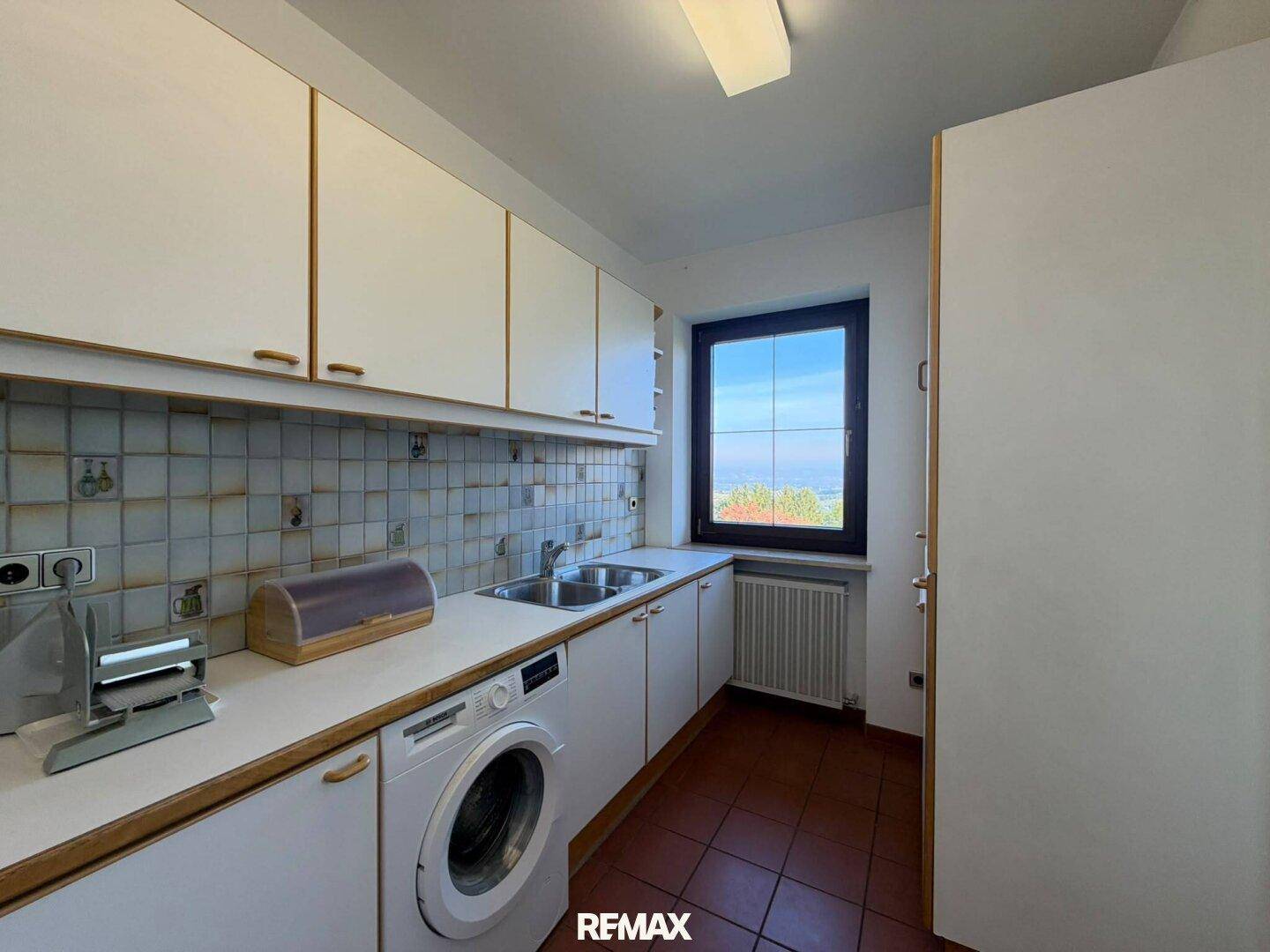 Funktionale Küche mit weißen Schränken, Waschmaschine und Fenster mit Fernblick.
