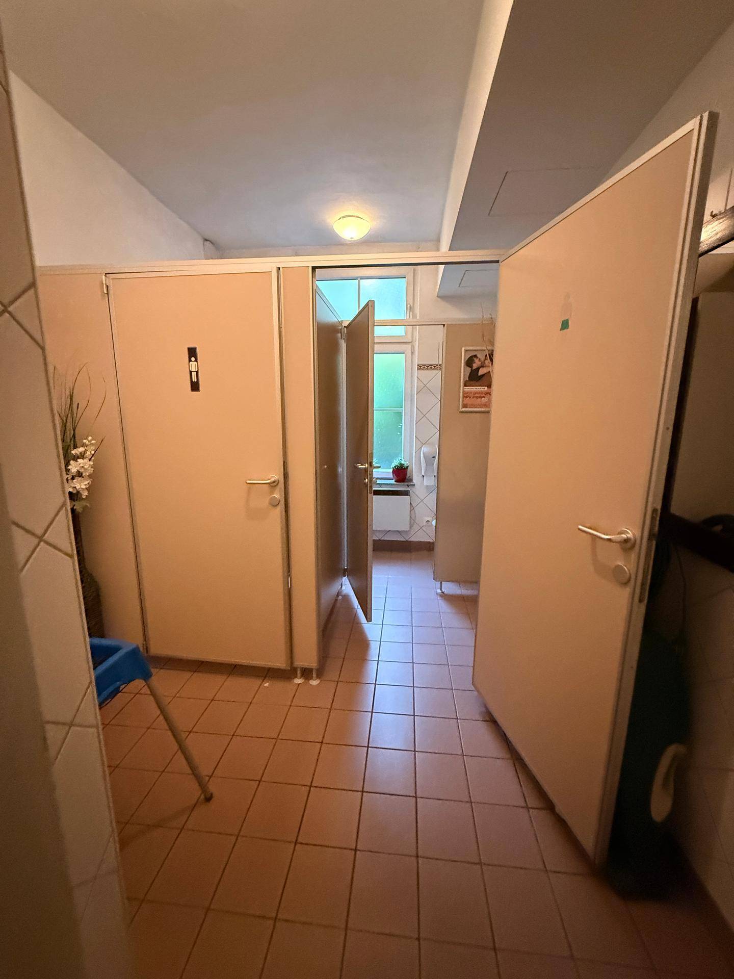 Flur zu den Toilettenkabinen in einem öffentlichen Bereich, mit hellen Fliesen und einer Tür zum Fenster.