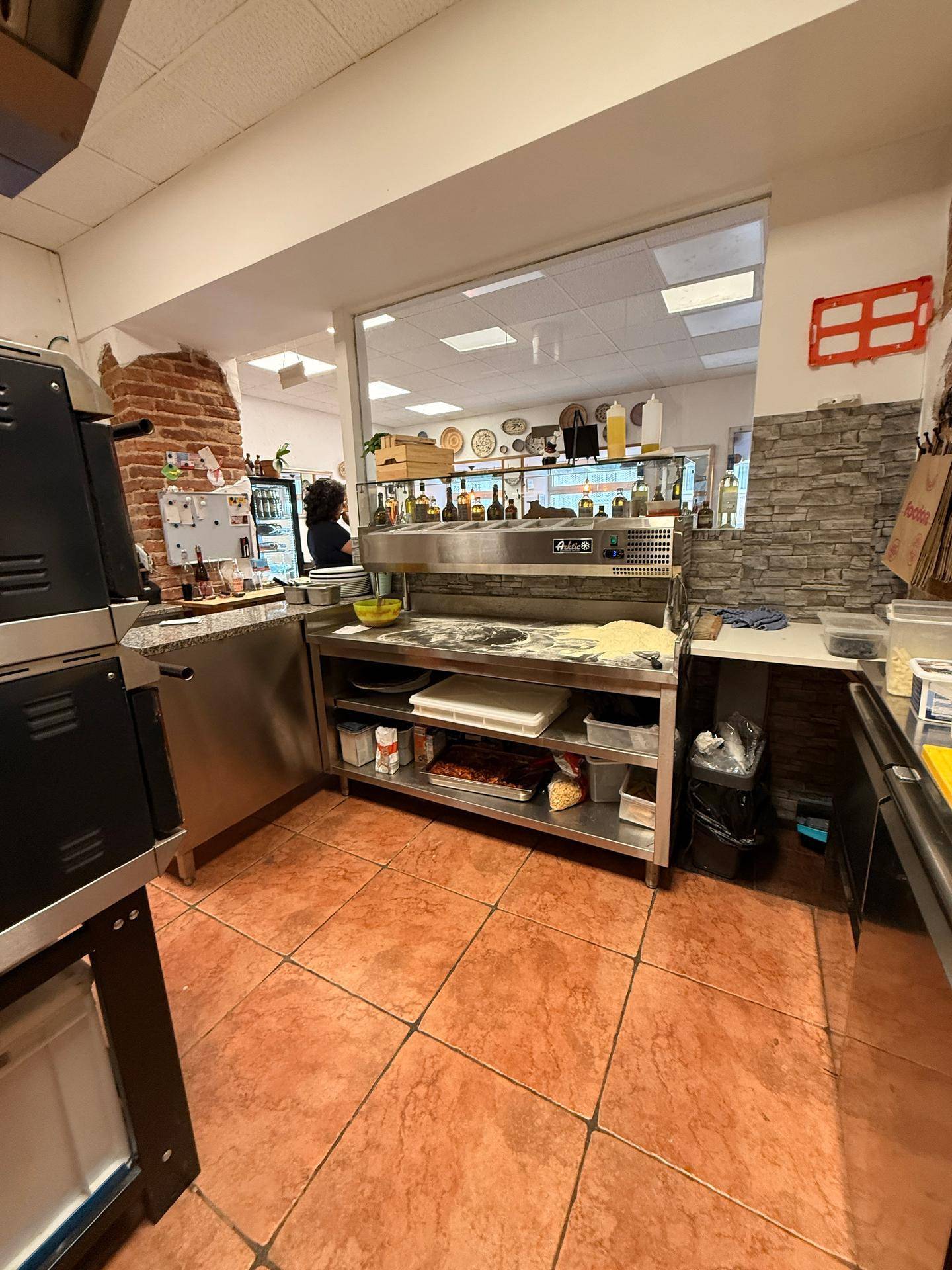 Gewerbliche Küche mit Durchreiche zum Gastraum, ausgestattet mit professionellen Pizzaöfen und Arbeitsflächen.