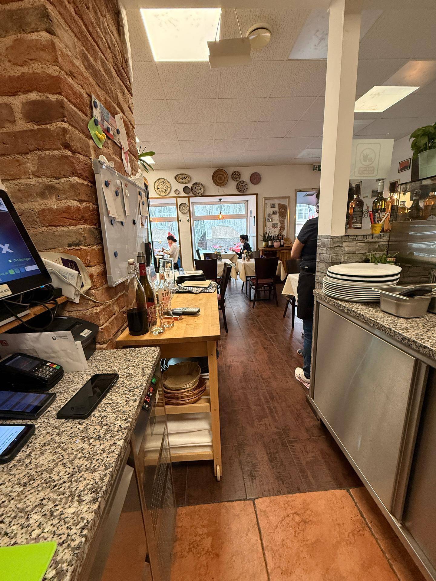 Blick vom Thekenbereich in den Gastraum eines Restaurants mit Tischen und Stühlen, beleuchtet durch Tageslicht.
